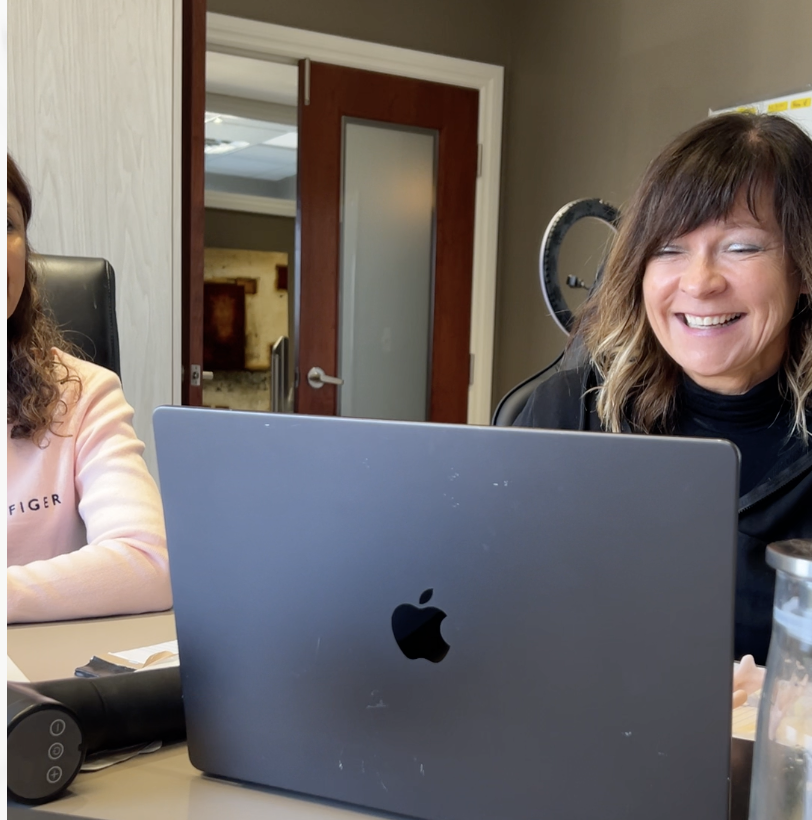 A woman with shoulder-length brown hair and a black top smiling while sitting at a table with a laptop in front of her.
