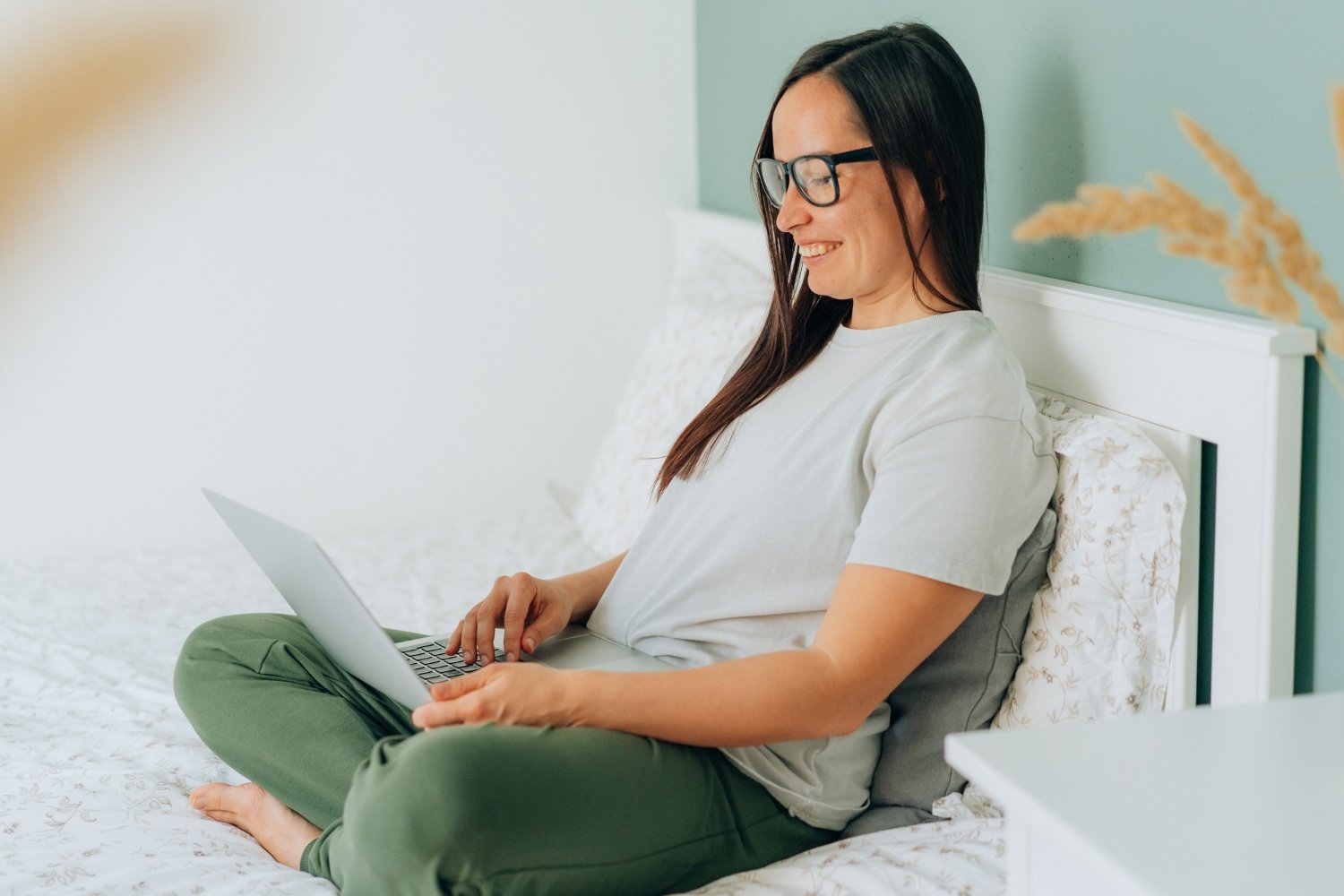 Frau sitzt auf dem Bett mit dem Laptop auf ihrem Schoß und lächelt.