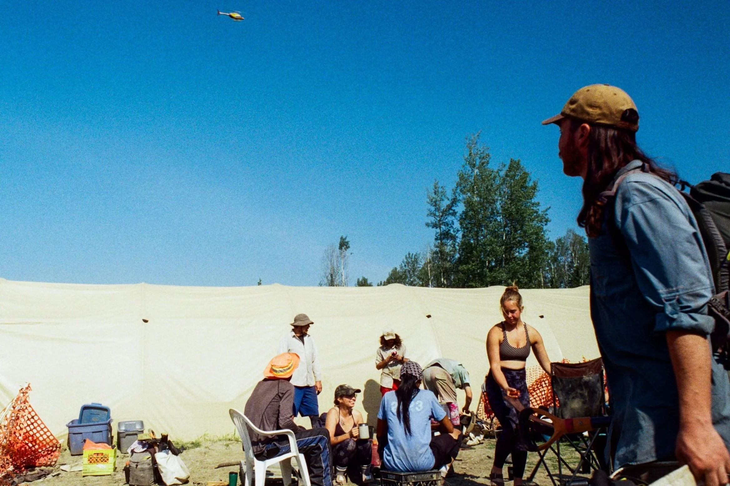 A group of people outdoors near a large beige tent, some seated and some standing, with a small helicopter flying in a clear blue sky.
