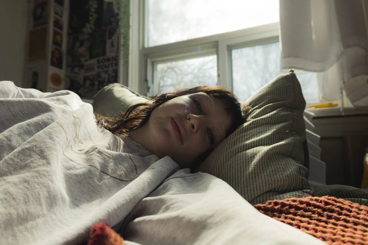 Young woman lying on bed with sunlight coming through a window, smiling with her eyes closed.