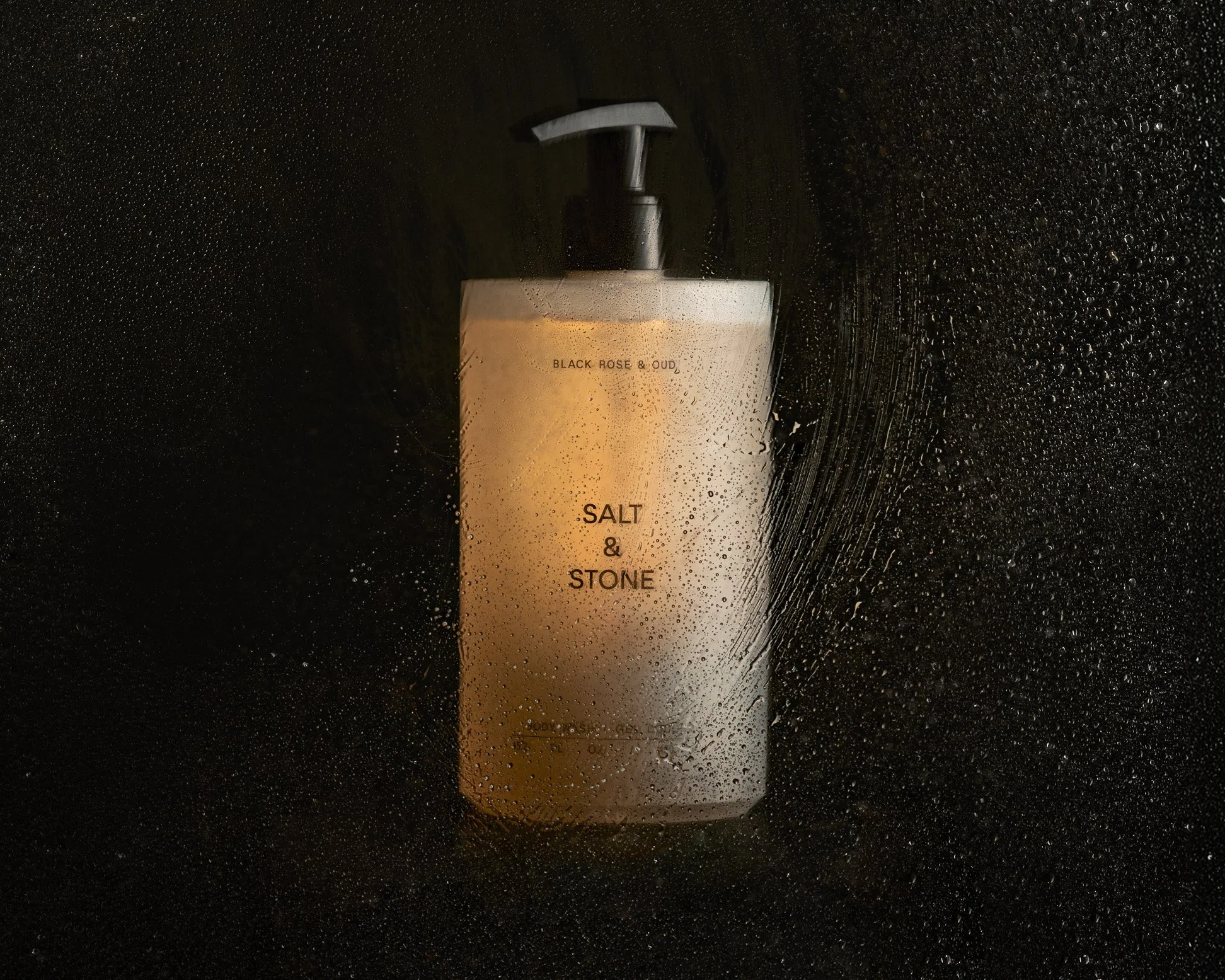 A bottle of Body Wash labeled 'Salt & Stone' with a pump dispenser, surrounded by water droplets on a black surface