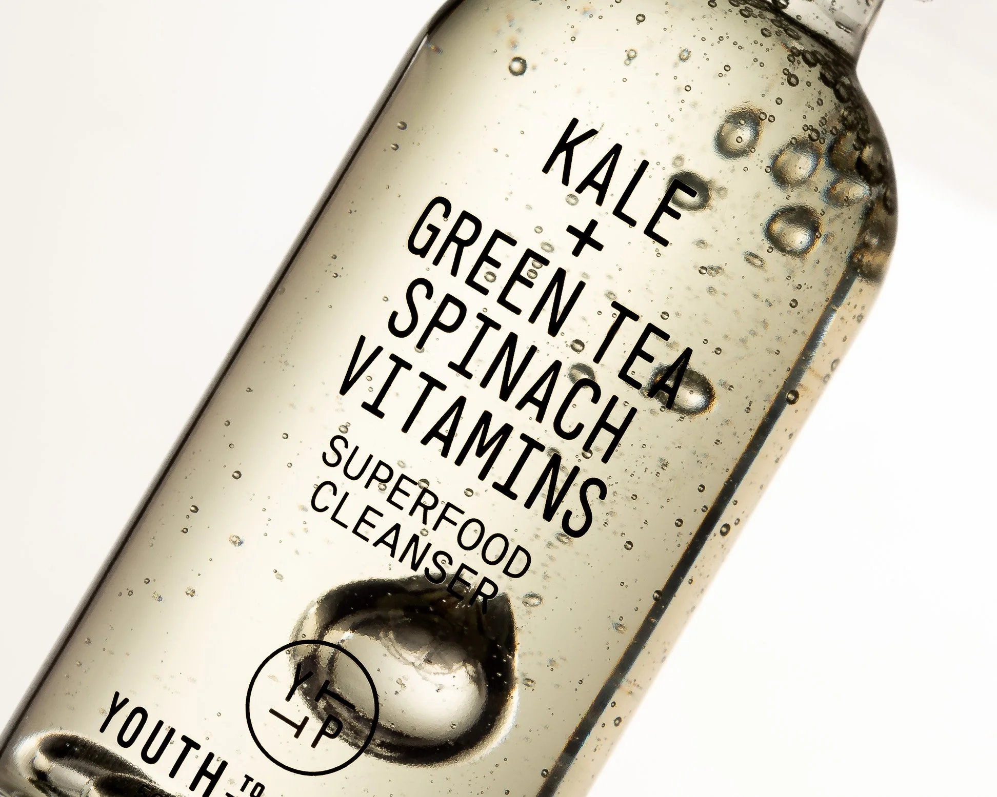 Clear skincare serum bottle with black text listing ingredients: kale, green tea, spinach, vitamins, superfood cleanser, with bubbles inside the bottle.