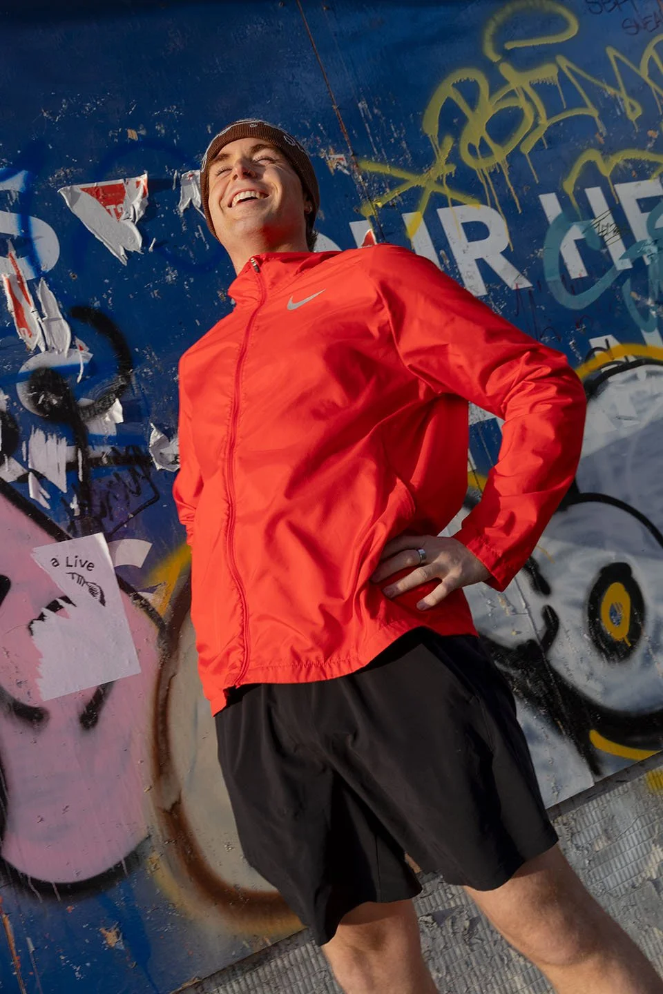 A young man wearing a red Nike windbreaker and black shorts standing in front of a graffiti-covered wall, smiling with his head tilted back.