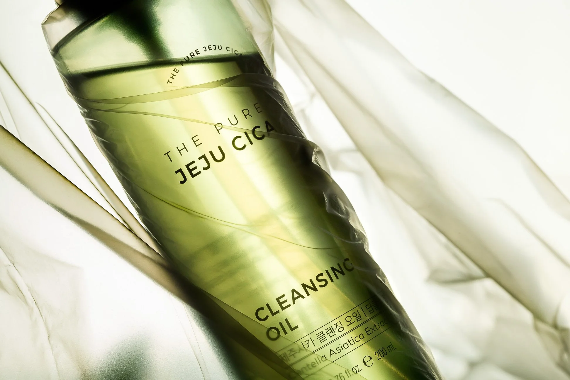A green bottle of The Pure Jeju Cicada Cleansing Oil wrapped in transparent plastic with white fabric background.