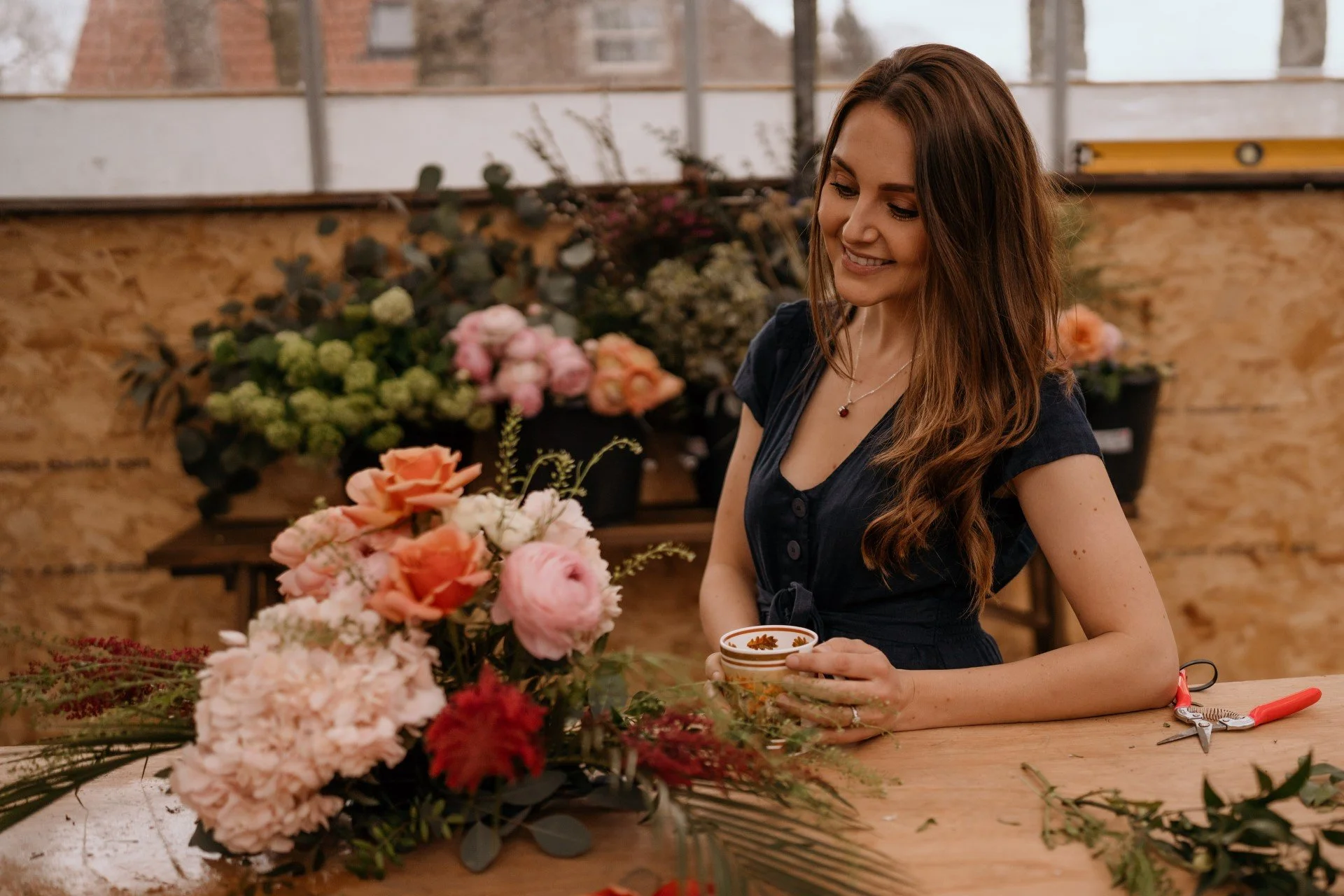 A woman with long wavy brown hair wearing a navy blue dress, holding a cup and smiling while working on floral arrangements in a workshop with various flowers and tools on the table.