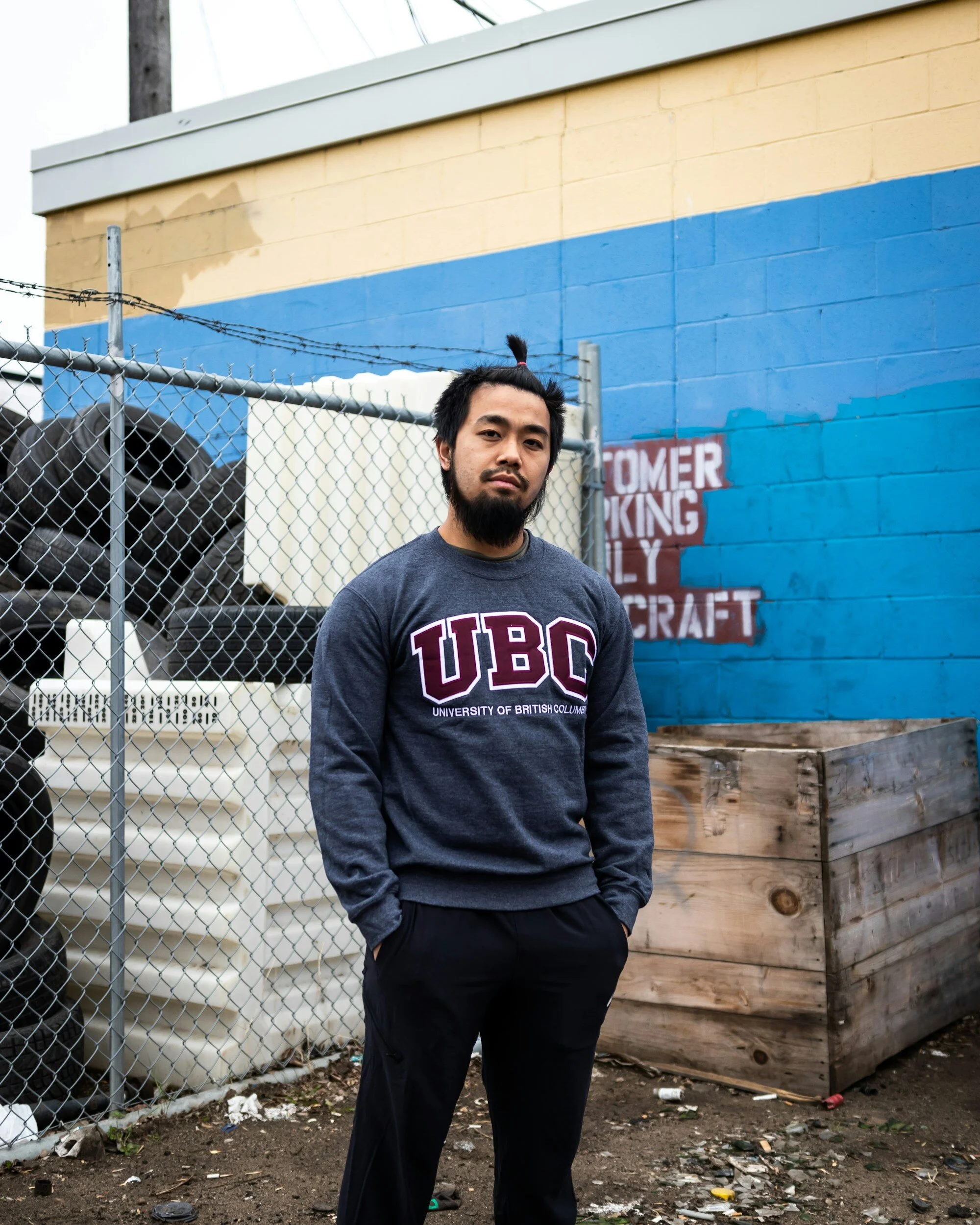 A man standing outdoors in front of a chain-link fence and a colorful wall with a painted sign. He is wearing a dark gray sweatshirt with 'UBC' and 'University of British Columbia' printed on it. The ground is dirt with some trash, and there is a large wooden planter box to his right.