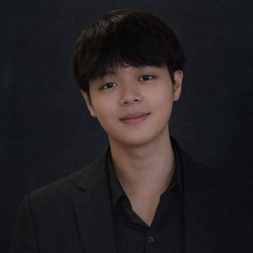 A young man with black hair, wearing a black shirt and blazer, posing against a dark background.