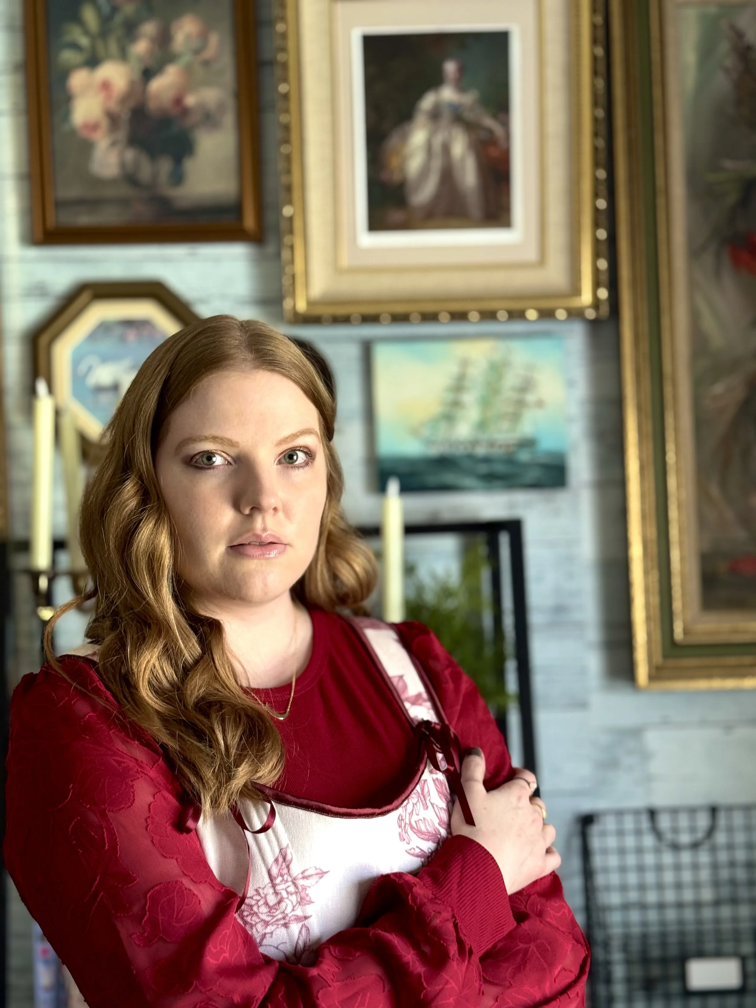 white woman with red hair, she is wearing a red one sleeve shirt with white accents. Her arms are folded over her chest, and she is standing in front of a wall of framed photos