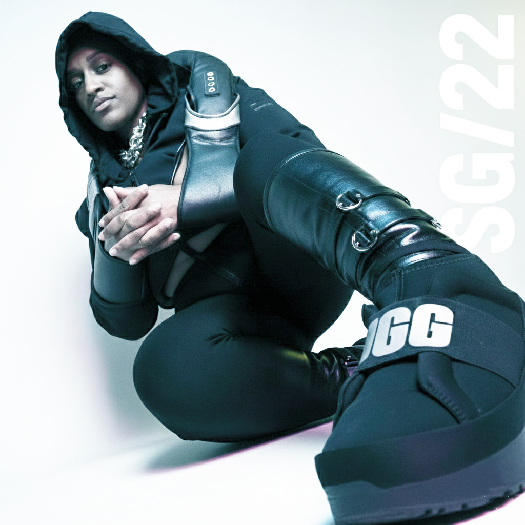 Model in a black hooded outfit and chunky platform boots seated against a pale background, with one boot close to the camera and SG/22 lettering alongside.