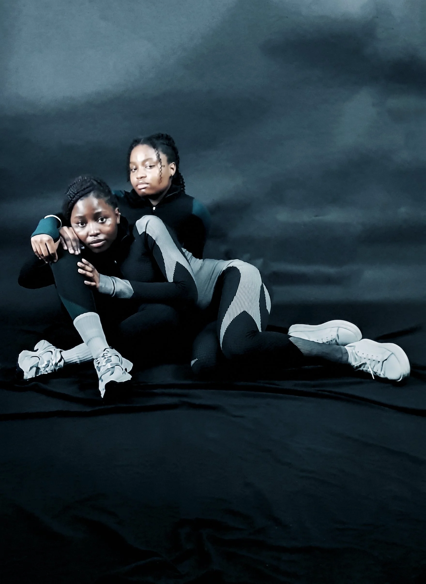Two models in black and grey fitted sportswear and white trainers posing closely together on a dark fabric backdrop.