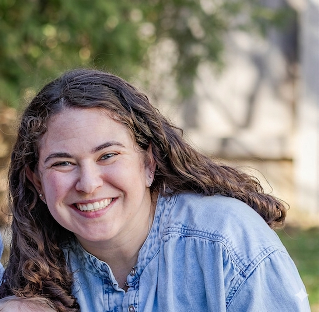 Close-up of a smiling woman with curly brown hair, wearing a light blue denim shirt, outdoors with greenery in the background.