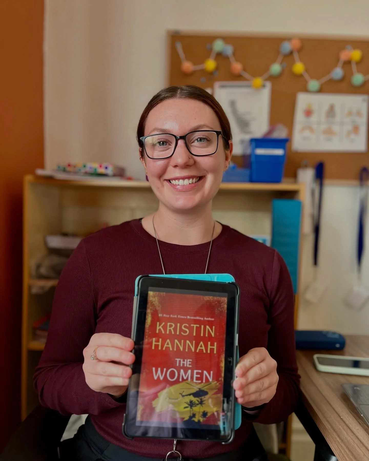 A smiling woman with glasses holding a Kindle e-reader displaying a book titled 'The Women' by Kristin Hannah in a room with a wooden desk and shelves, a bulletin board with colorful pins, and papers and electronic devices in the background.