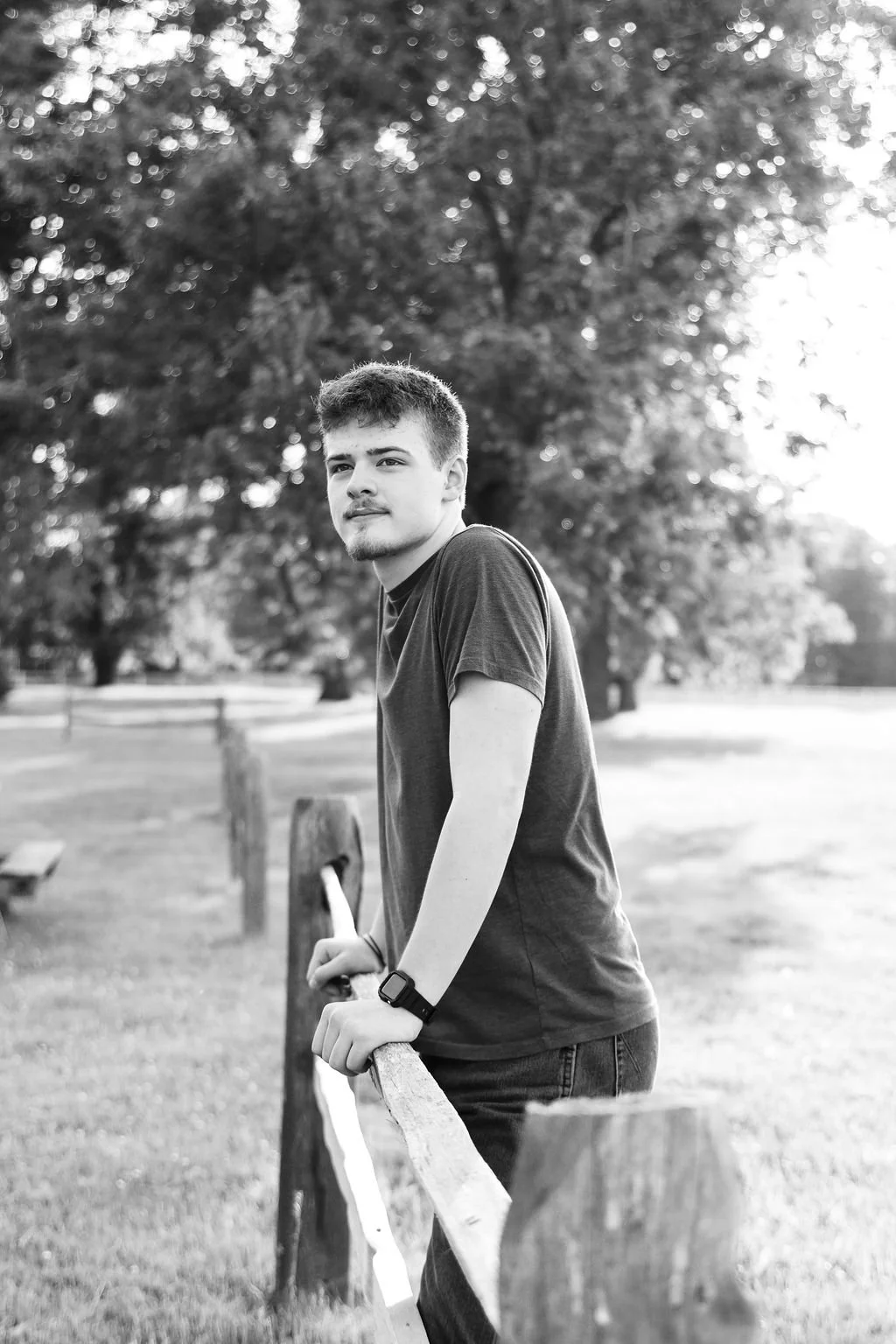 A young man leaning on a wooden fence in a park, looking into the distance with a thoughtful expression, during sunset. Black and white photo.