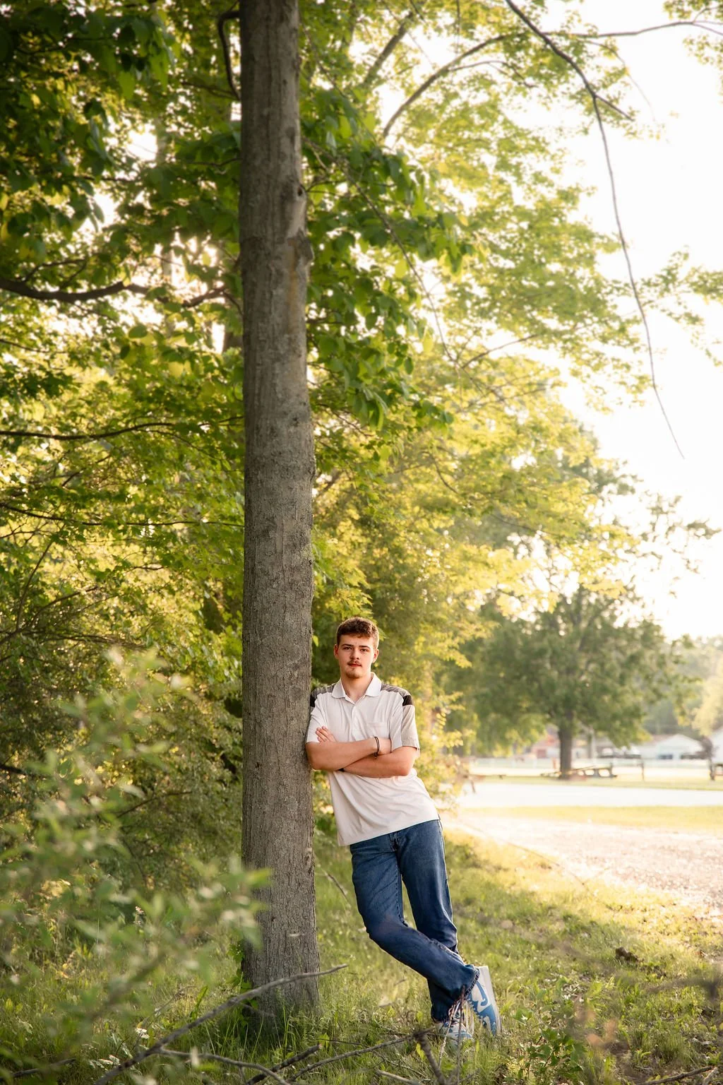 A young man with a mustache and short hairstyle leaning against a tree with green foliage, arms crossed, in a park during late afternoon or early evening.