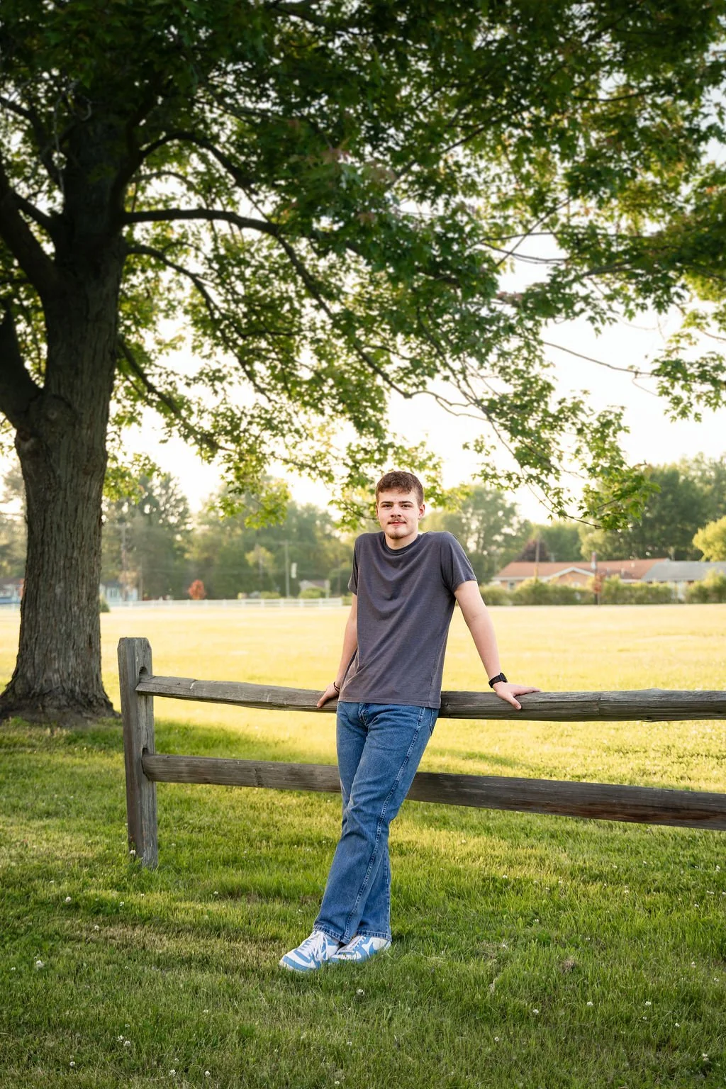 A young man standing outdoors beside a wooden fence under a large leafy tree in a grassy park area, with houses visible in the background.