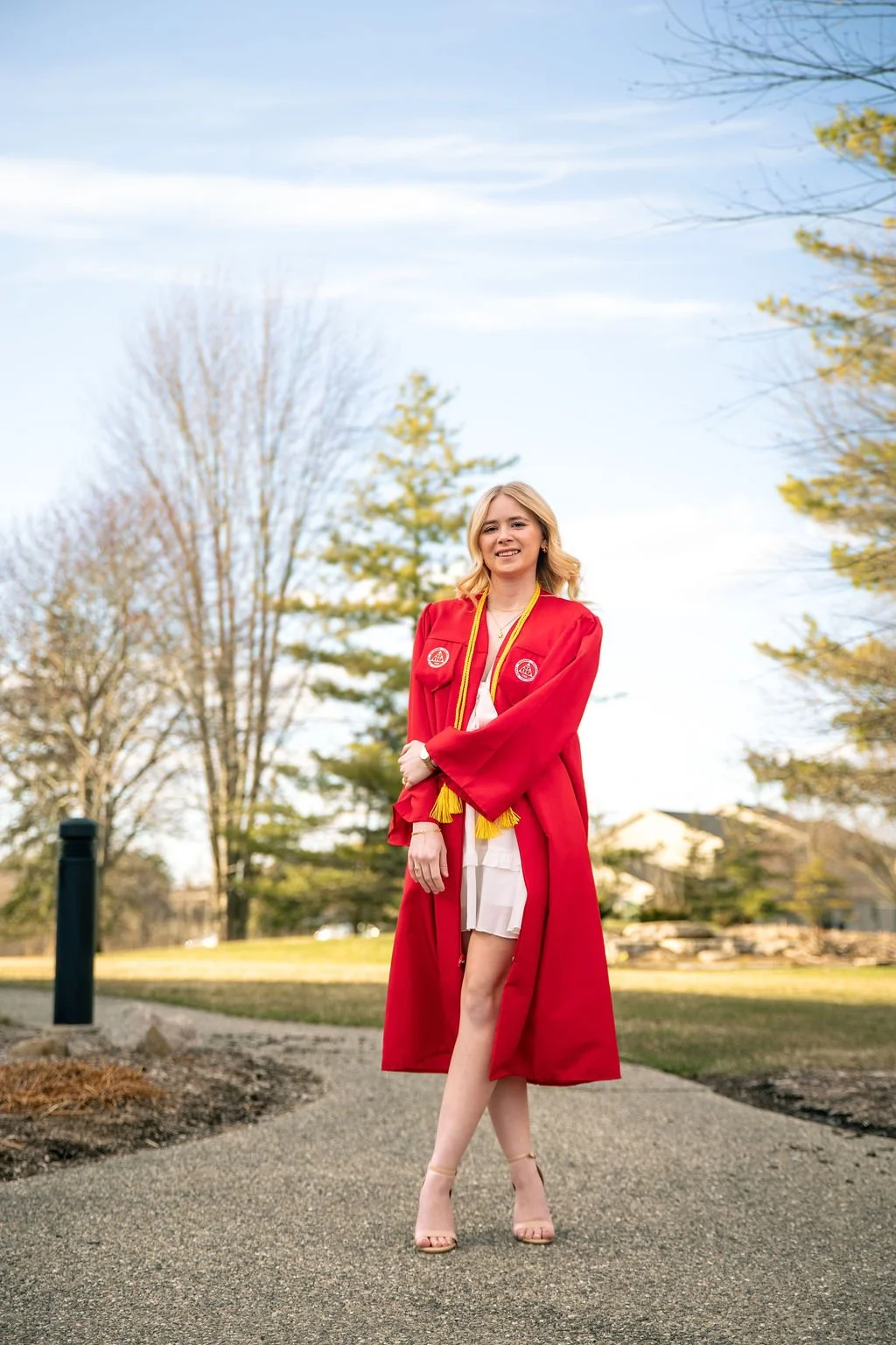 A young woman in a red graduation gown and tassel standing outside on a paved path, smiling during daytime with trees and a blue sky in the background.