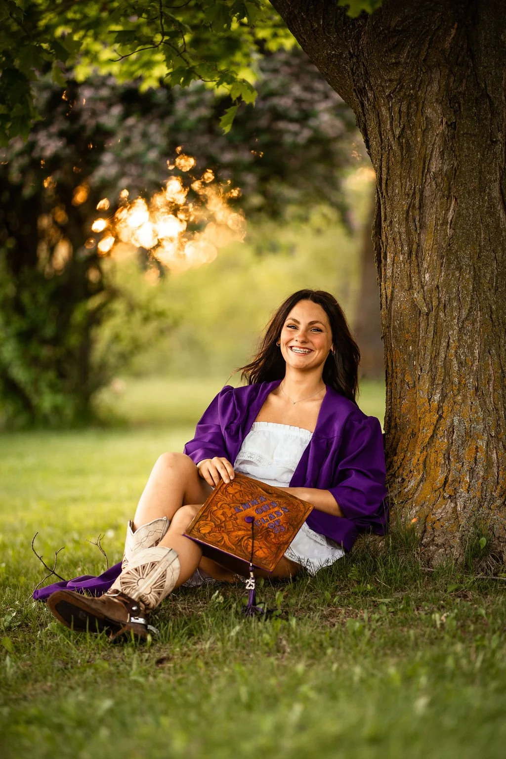 A young woman wearing a purple graduation gown sitting on the grass next to a large tree, holding a decorated graduation cap, smiling happily outdoors during sunset.