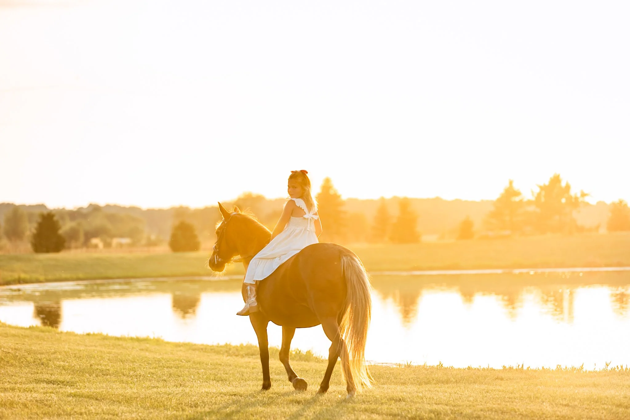 A young girl in a white dress riding a brown horse near a lake during sunset.