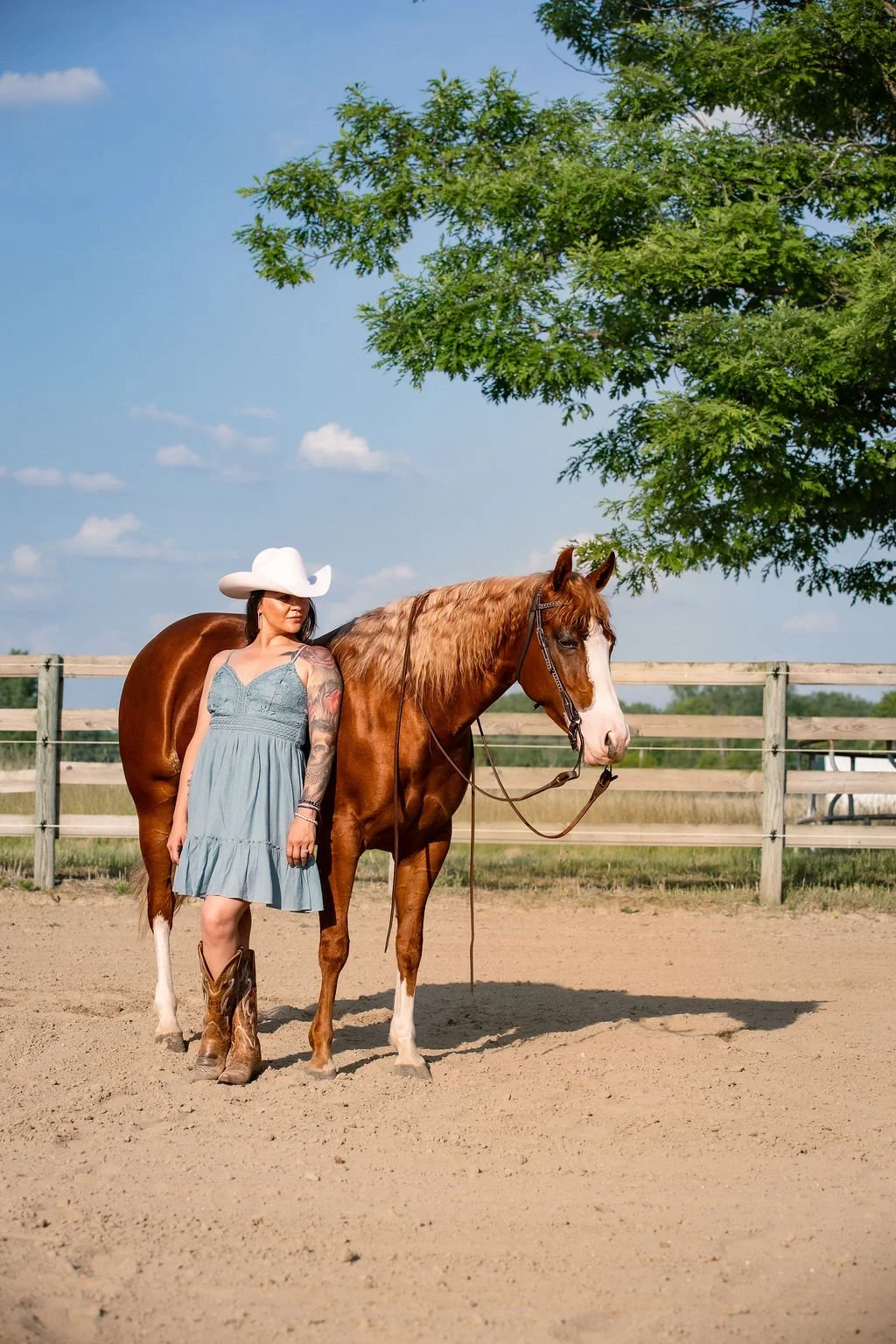 A woman in a light blue dress, cowboy boots, and a white cowboy hat standing next to a chestnut and white horse in a fenced outdoor area, with a large green tree and a blue sky with scattered clouds in the background.