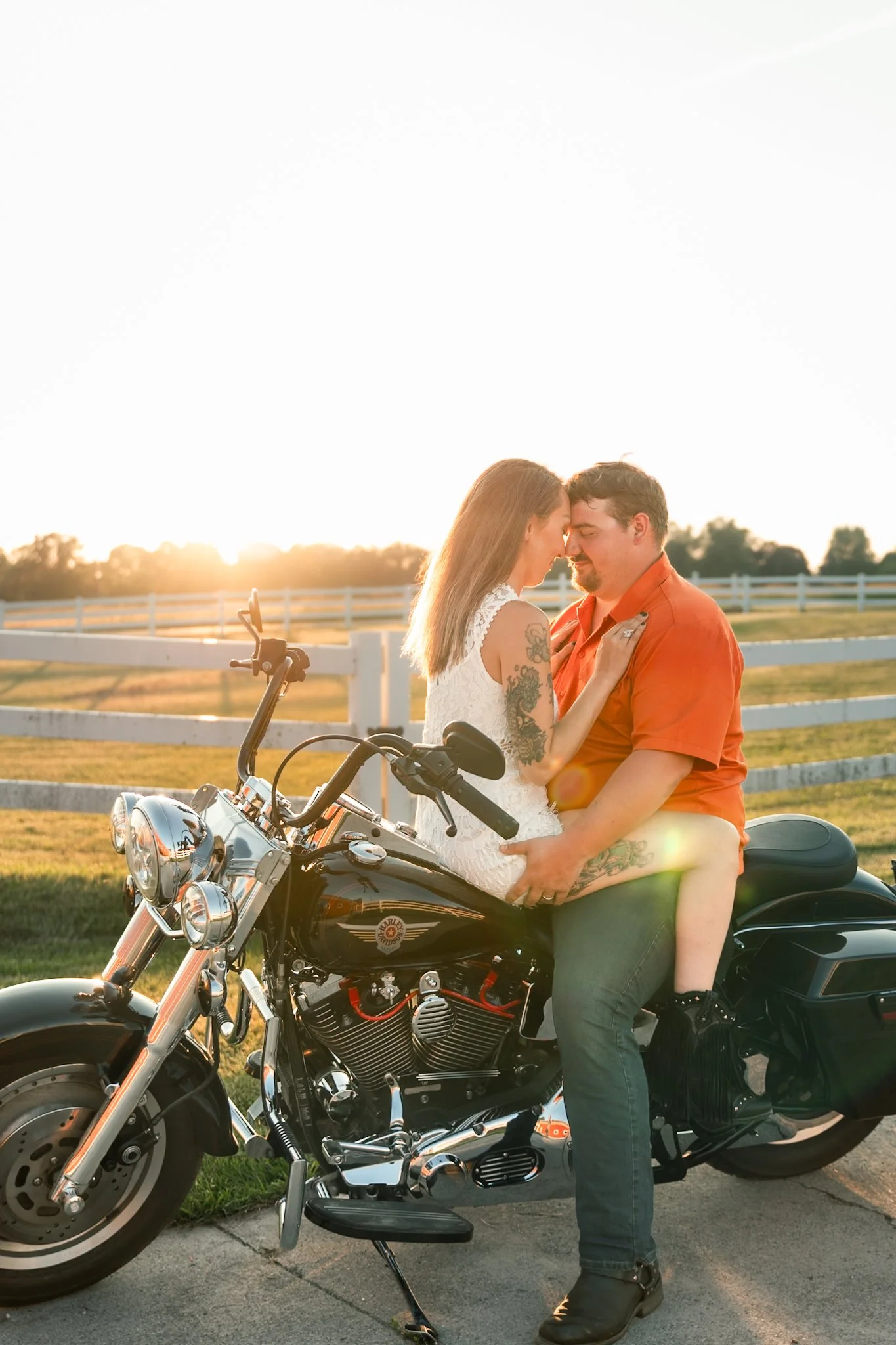 A couple sitting closely on a black motorcycle outdoors during sunset, with a white fence and open field in the background.