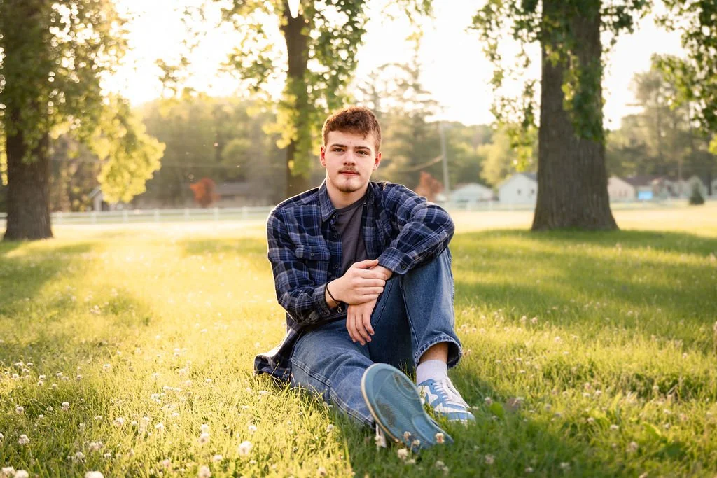 A young man sitting on green grass in a park with trees in the background during sunset.