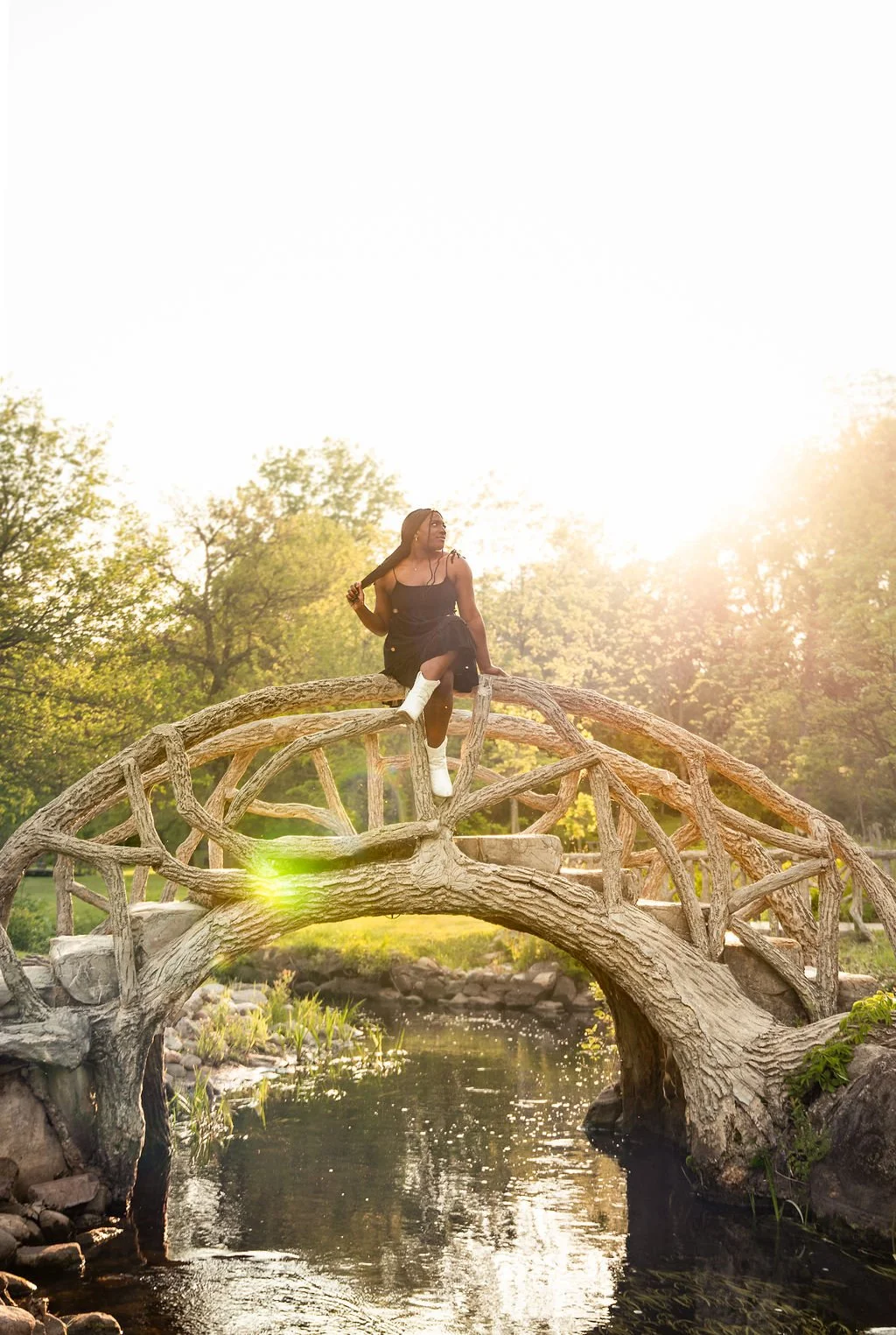A woman sitting on a twisted wooden bridge over a small river in a park during sunset.