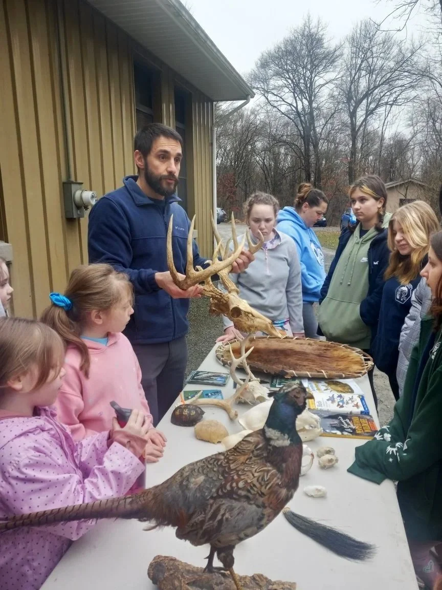 A man holding a deer skull with antlers shows it to a group of children gathered around a table with various animals and bones in an outdoor setting.