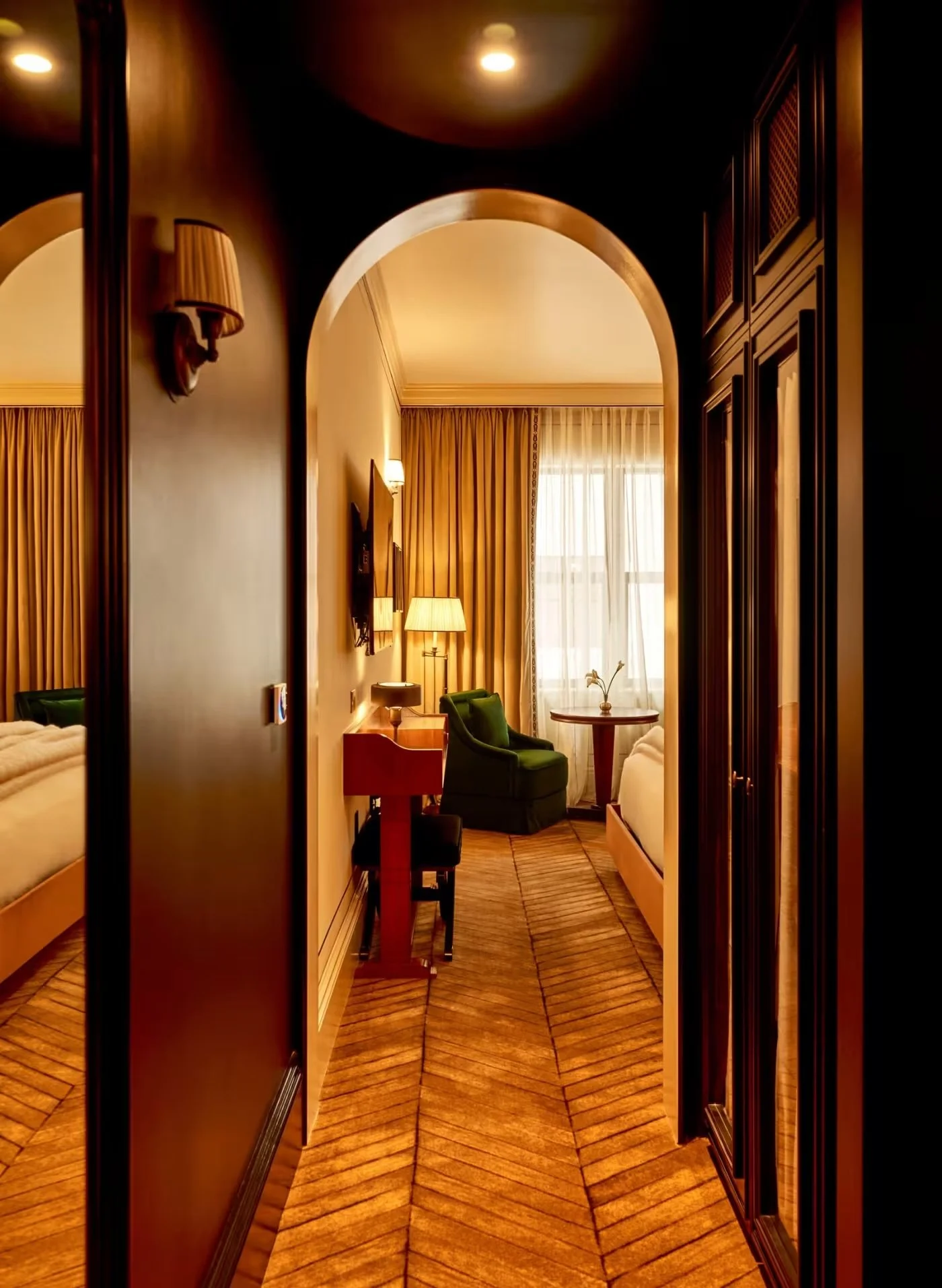 View through an arched doorway into a hotel room with a bed, green armchair, small round table with a flower, and curtains.