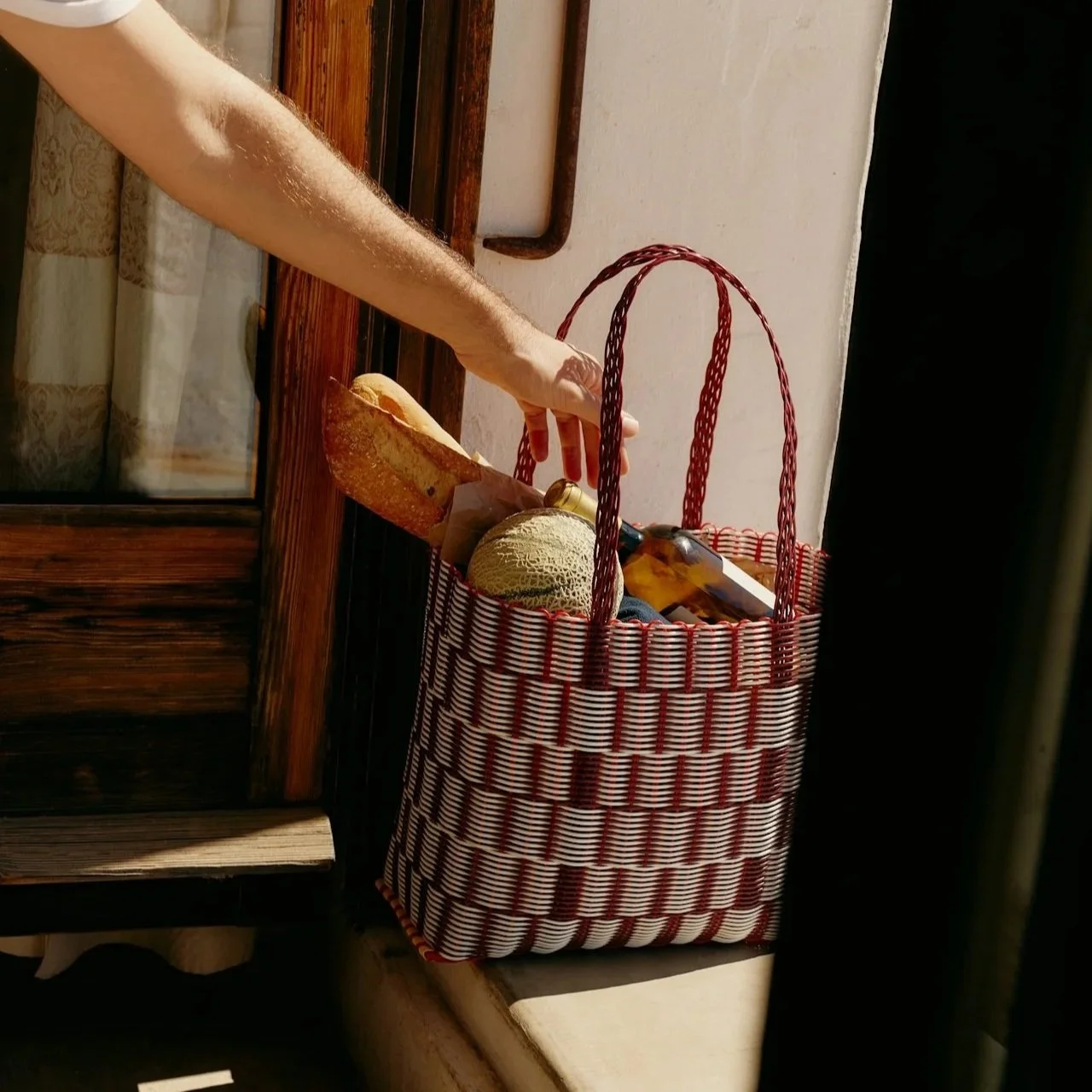 Red woven basket filled with various items on a windowsill, partially obscured by a person's arm reaching in from the left.