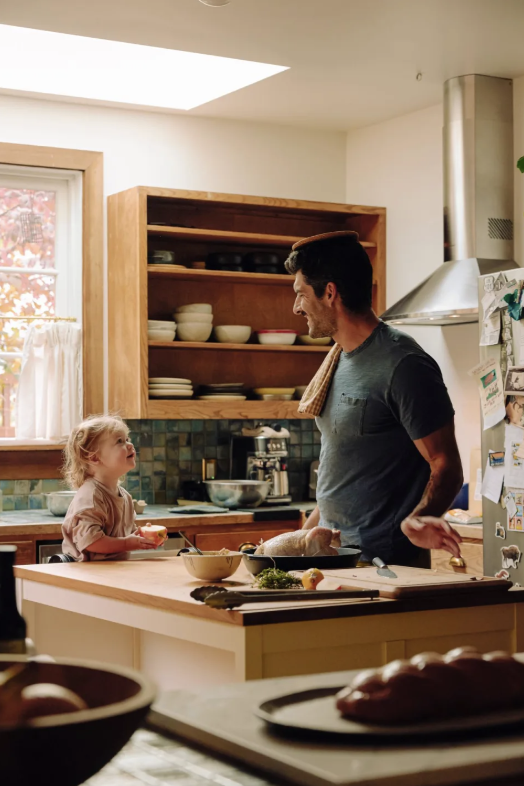 A man and a young girl are smiling and talking in a kitchen, with the man standing near a counter with a roasted chicken. The girl is holding a piece of food, and there are bowls, a cutting board with vegetables, and a window with curtains in the background.