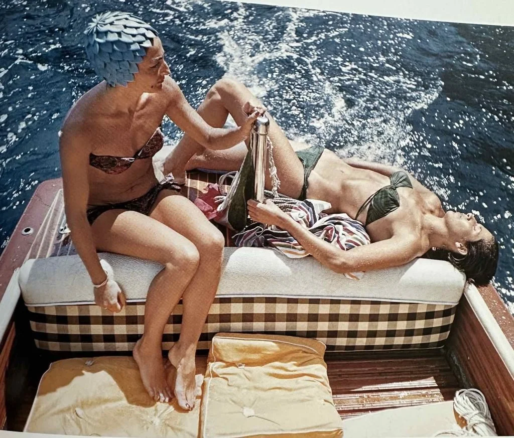 Two women in swimsuits relaxing on a boat, one lying down with a towel, the other sitting on the edge, holding a bottle, with water visible in the background.