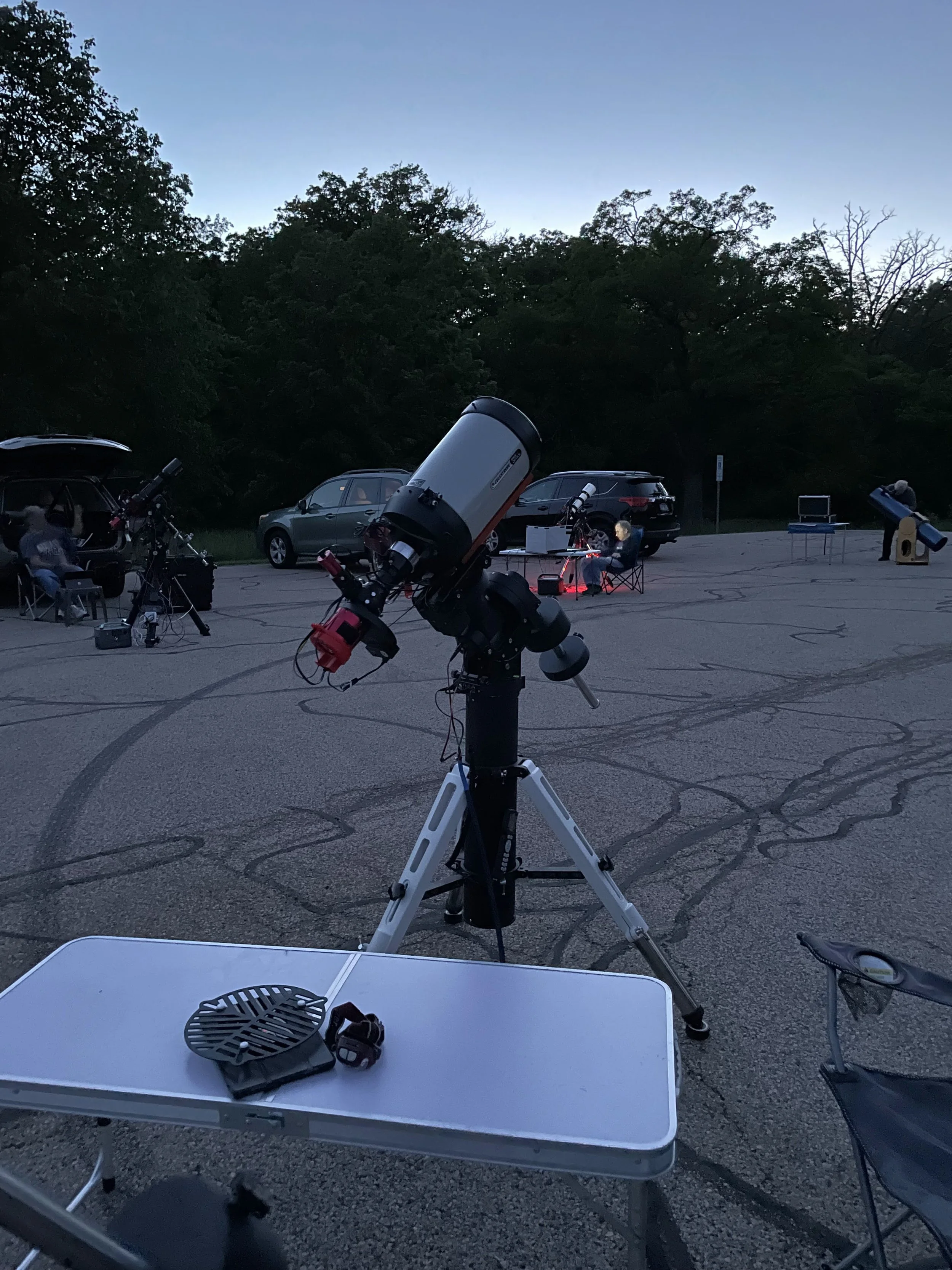 Outdoor stargazing event with telescopes on a parking lot, surrounded by trees and cars. People are seated nearby using telescopes in the evening.