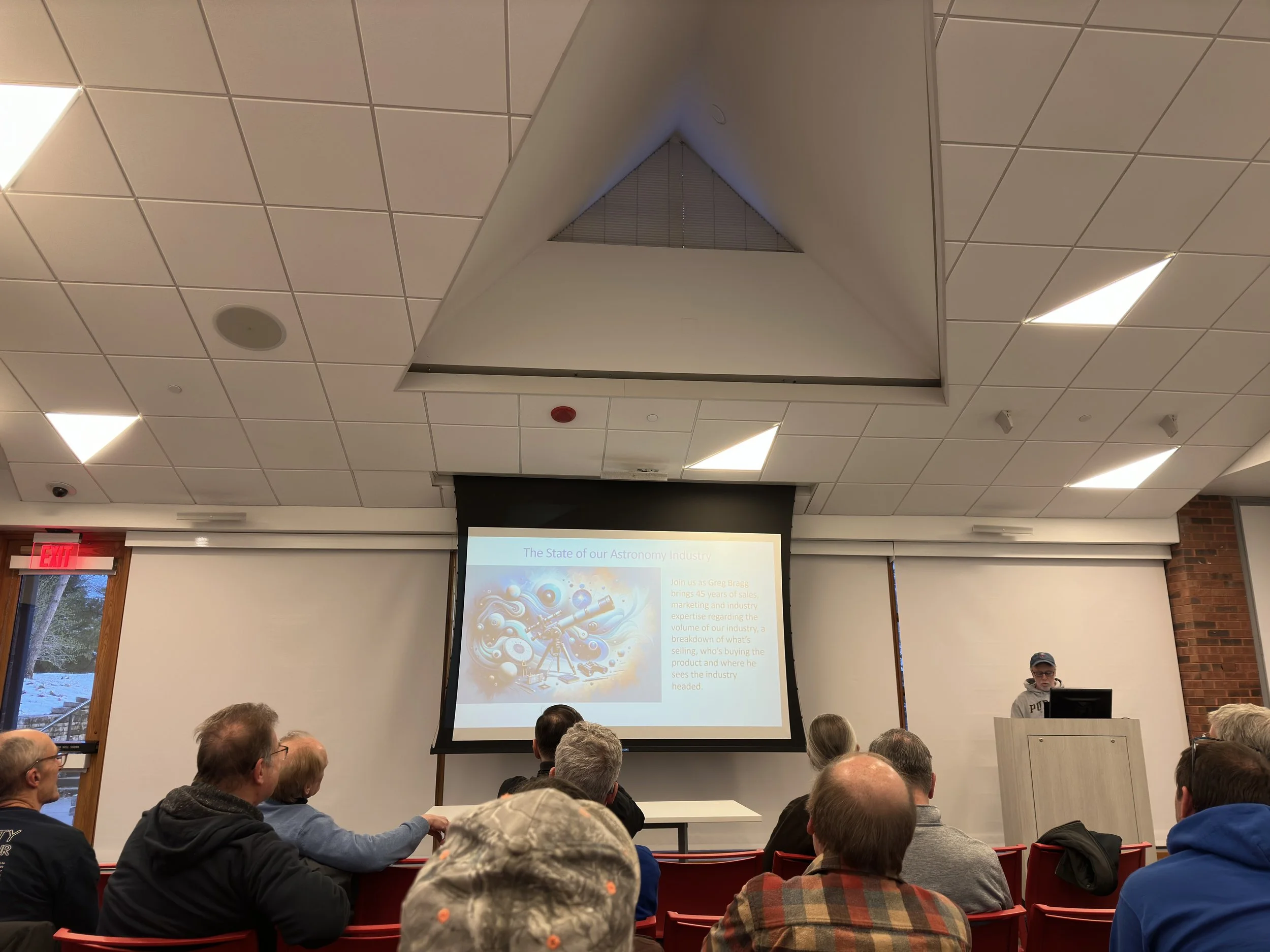 Indoor presentation in conference room with a speaker at the podium, slide on astronomy industry, and audience seated.
