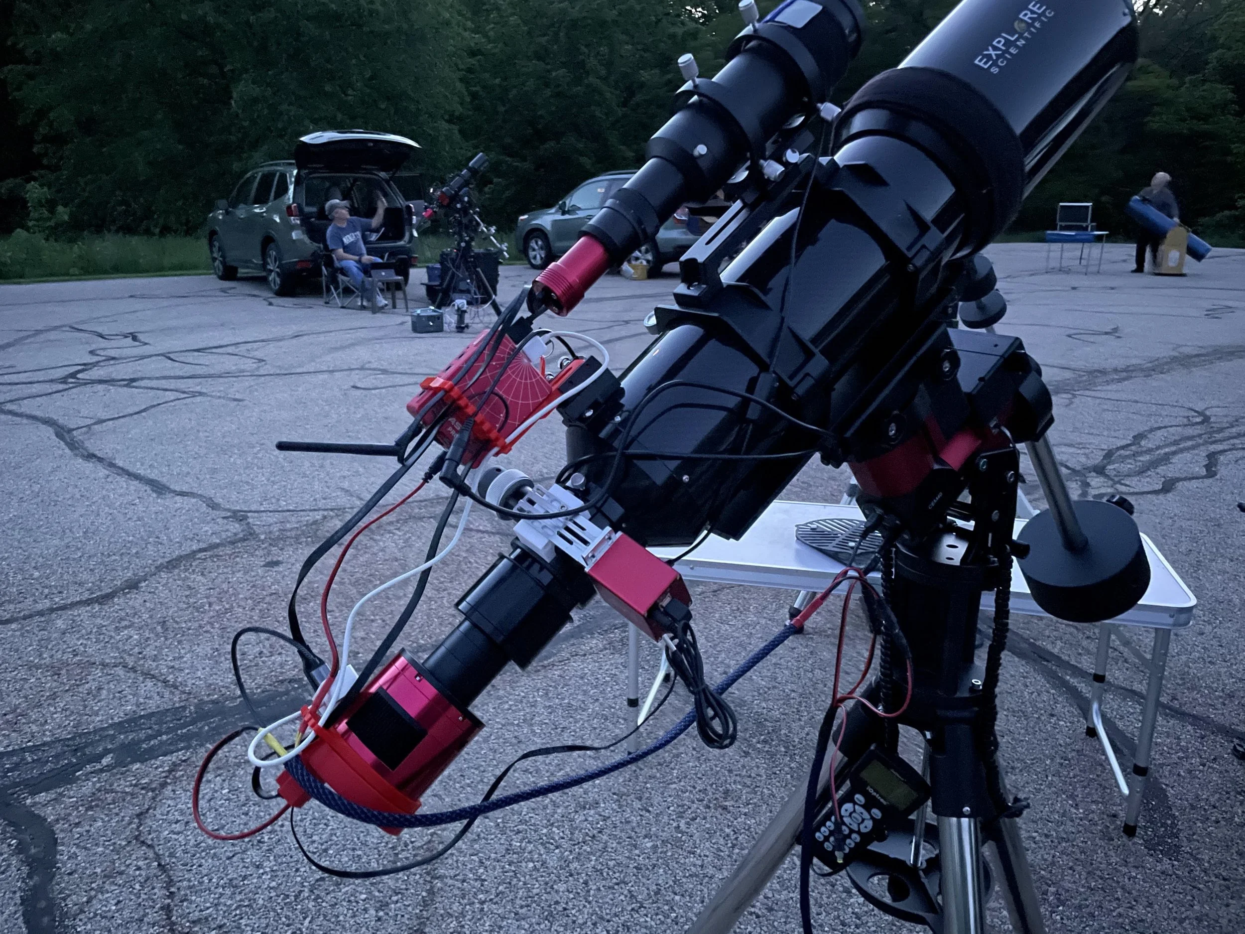 Close-up of a telescope setup with cables and equipment in a parking lot at dusk, with people and cars in the background.