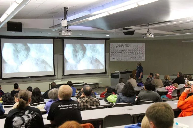 A classroom with students seated facing two large projection screens displaying a presentation. A teacher stands near the front. The periodic table of elements is on the wall.