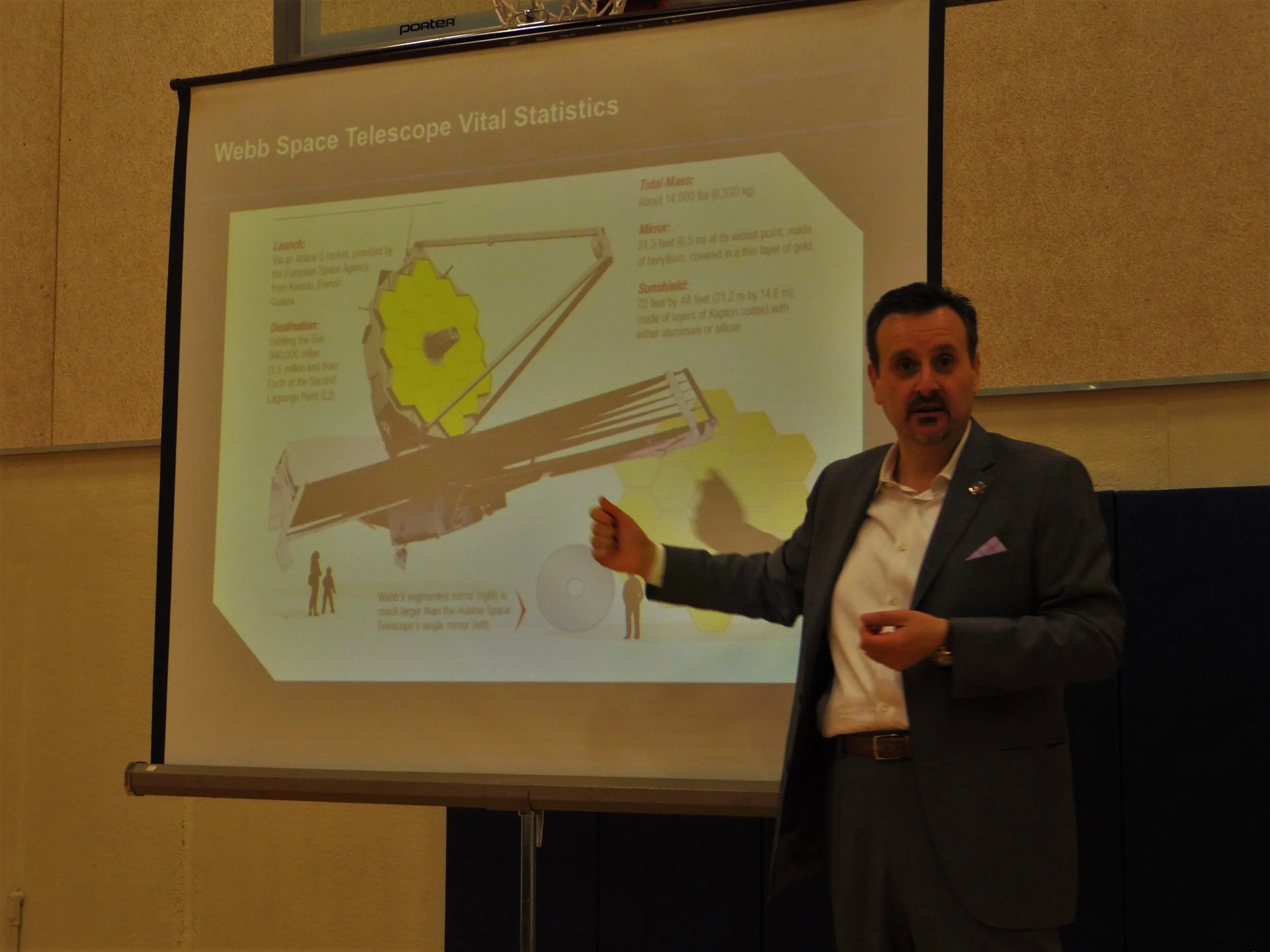 A man in a suit gives a presentation on the Webb Space Telescope, pointing to a slide with technical details and an illustration of the telescope.