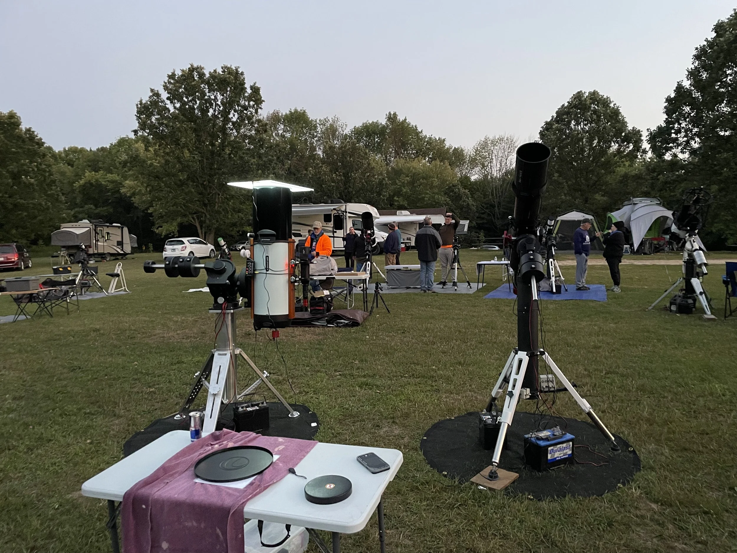 People setting up telescopes in a grassy area for stargazing event, surrounded by trees and parked cars.