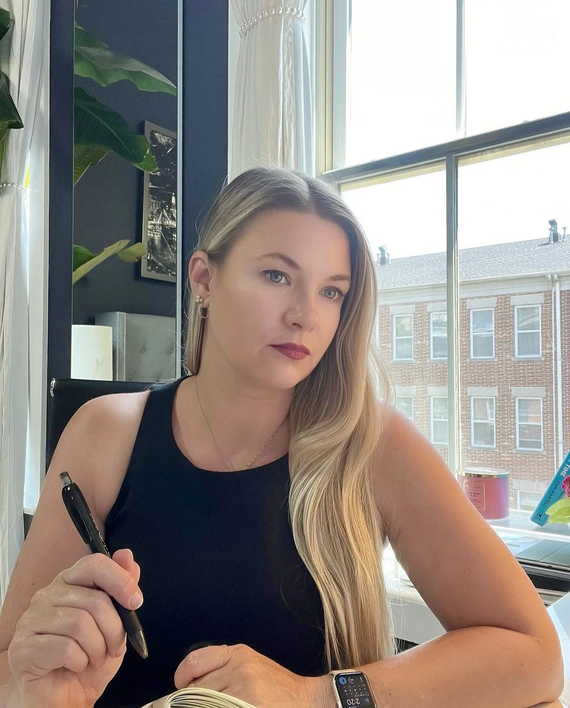 A woman with long blonde hair, wearing a black sleeveless top, sitting at a desk near a large window. She is holding a pen, with a serious expression on her face. There is a house visible outside the window, and indoor plants and decor are in the background.