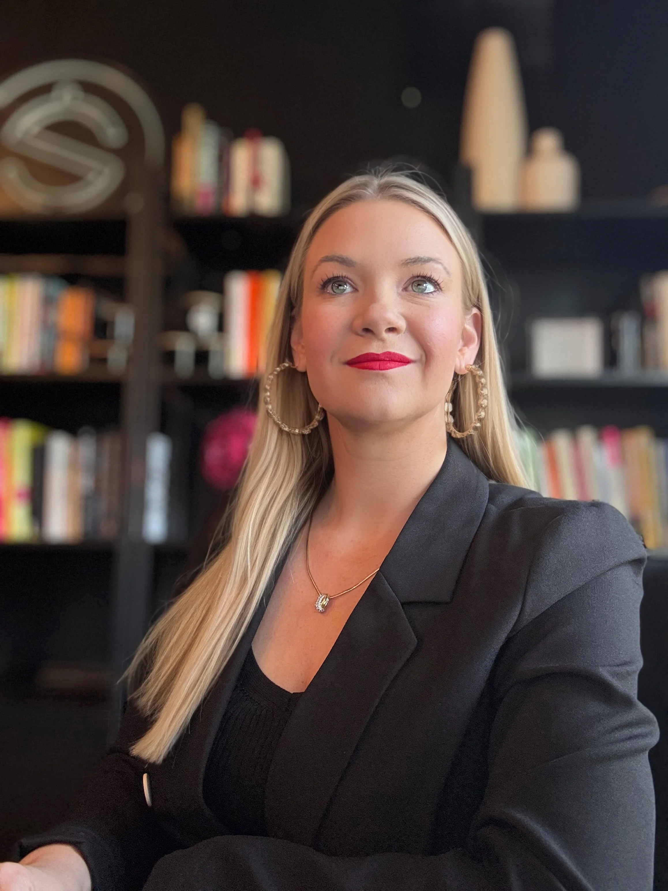 A woman with long blonde hair, red lipstick, and wearing gold hoop earrings and a black blazer, sitting in front of a blurred background with books on shelves.
