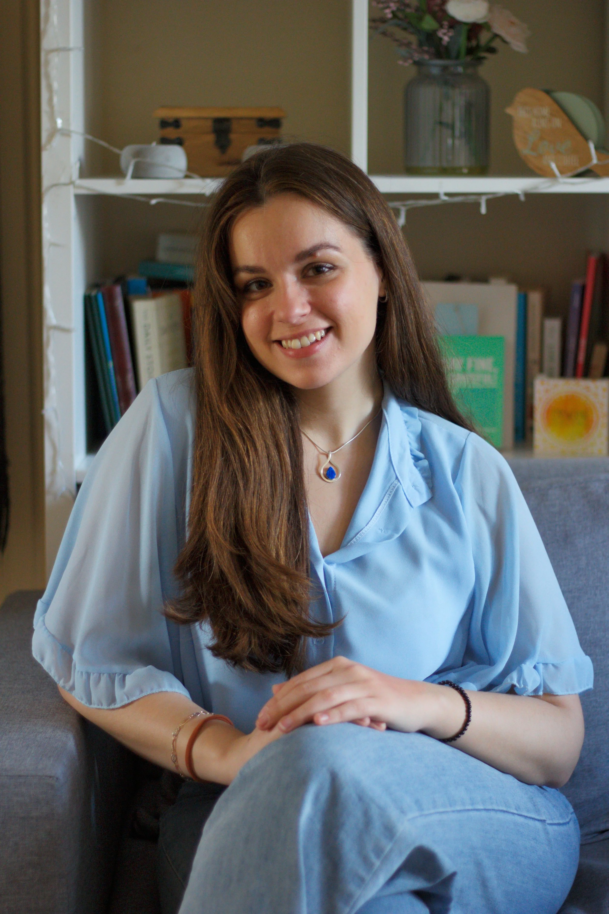 A young woman with long brown hair sitting on a grey couch, smiling at the camera in a cozy room with bookshelves and decorative items in the background.