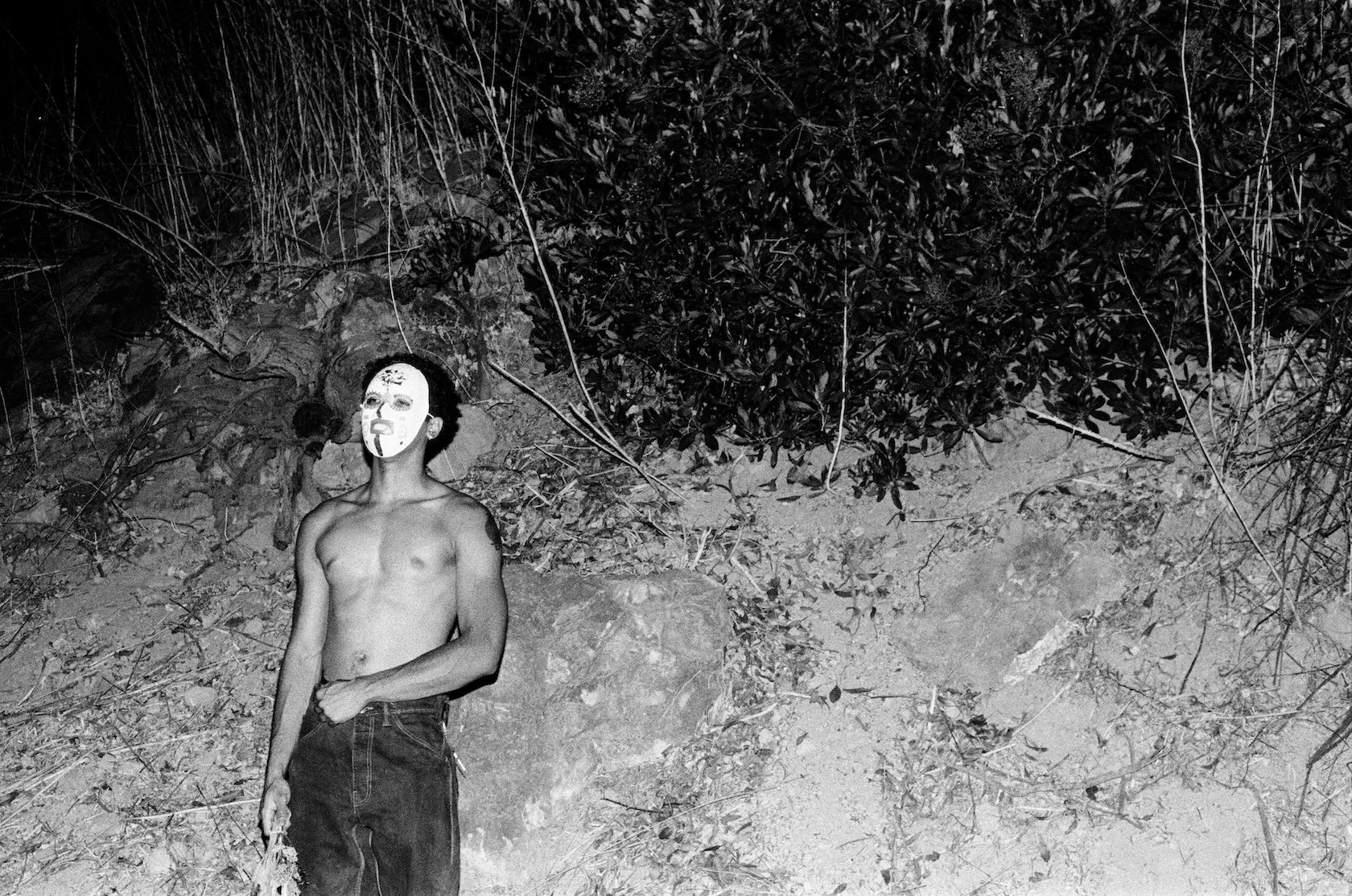 A shirtless young man wearing a painted mask stands outdoors at night beside a rocky hillside and dense brush; the black-and-white photograph is lit by flash, leaving much of the surrounding landscape in darkness.