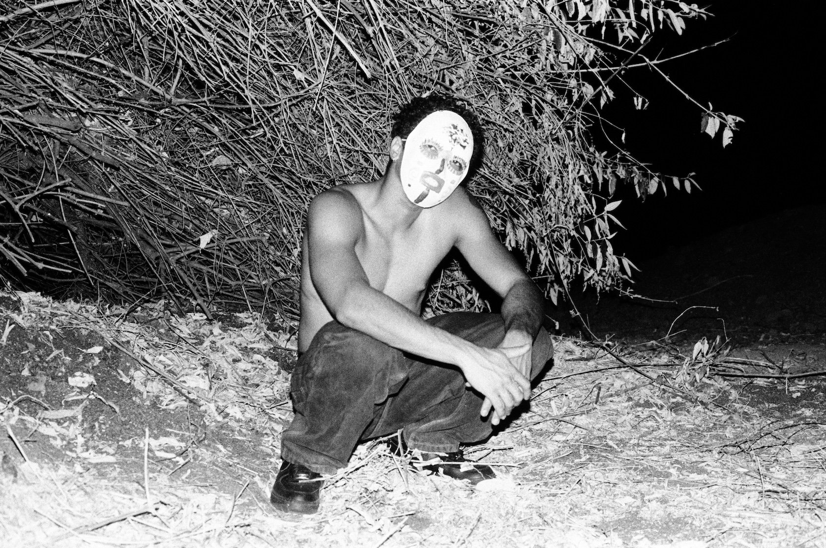 A shirtless young man wearing a painted mask sits low among dry branches and brush at night, resting his arms across his knees; the wide black-and-white image is illuminated by direct flash.