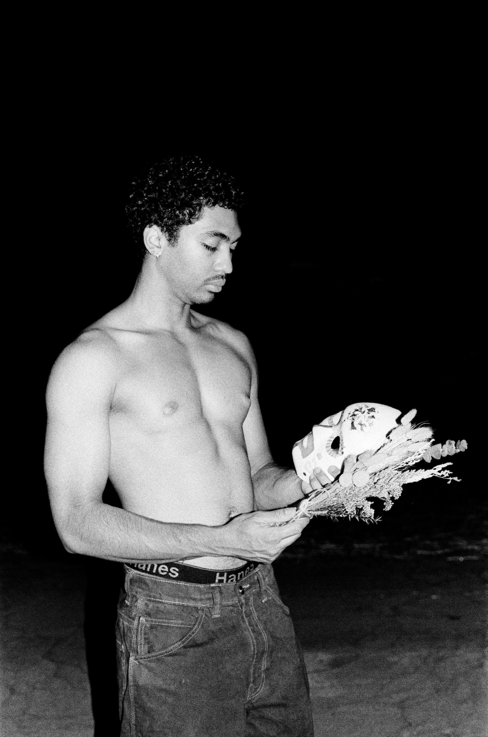 A shirtless young man stands against a dark nighttime background, looking down while holding a decorated painted mask and a small bundle of dried flowers in his hands; the black-and-white image is lit by direct flash.