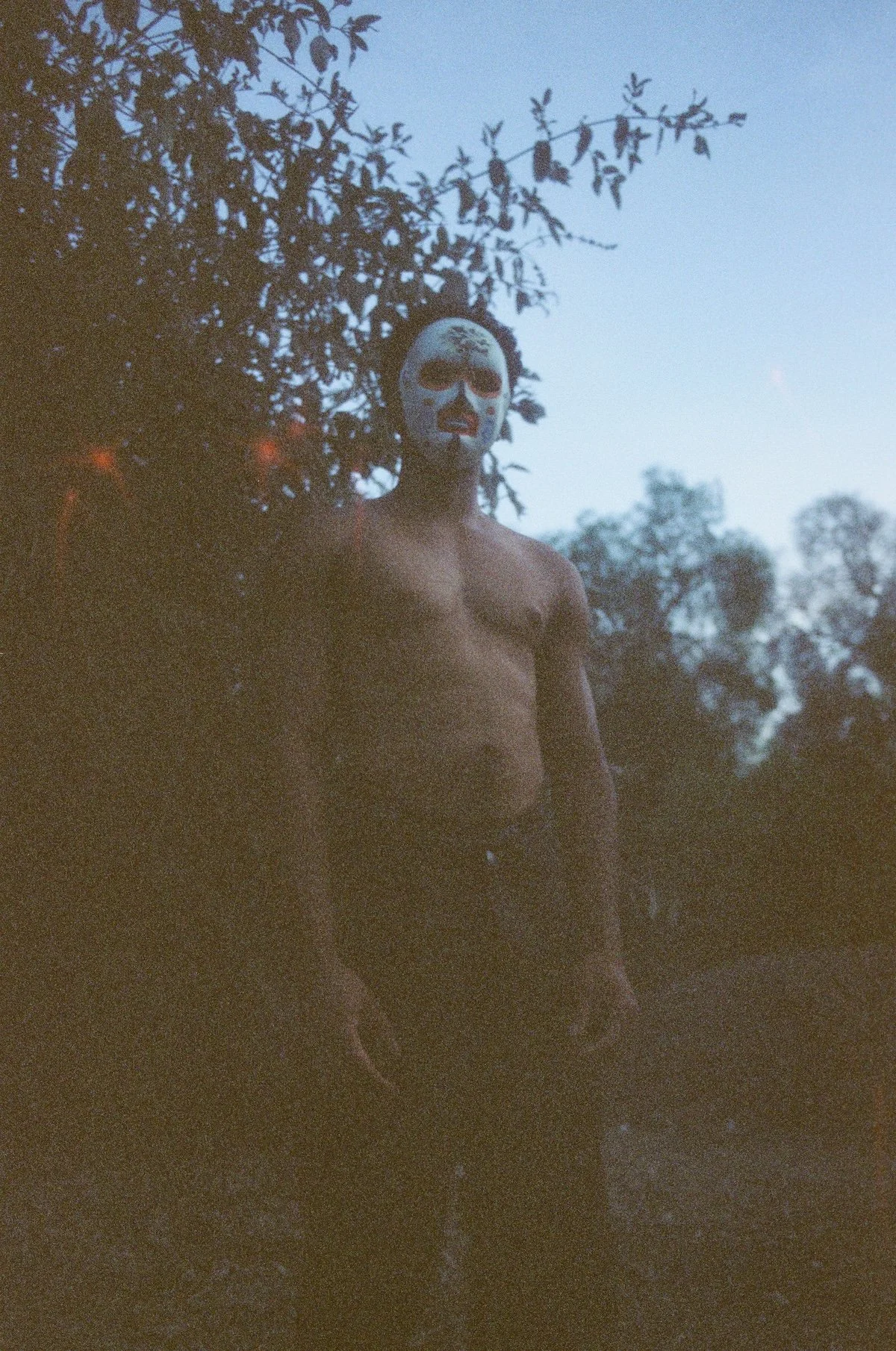 A shirtless young man wearing a pale painted mask stands outdoors at dusk beneath a blue-gray sky; the grainy color image is softly lit, with dark trees and brush surrounding him.
