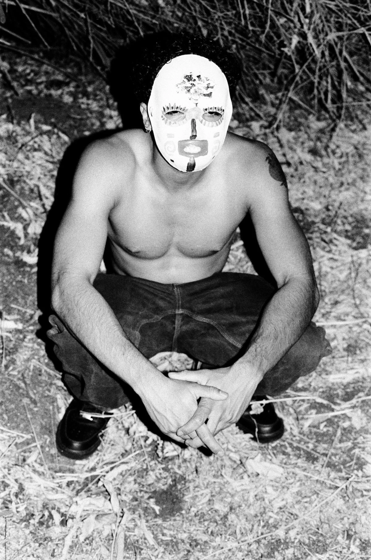 A shirtless young man wearing a painted mask crouches outdoors at night with his hands clasped in front of him; the black-and-white photograph is lit by direct flash against dry brush and leaves.