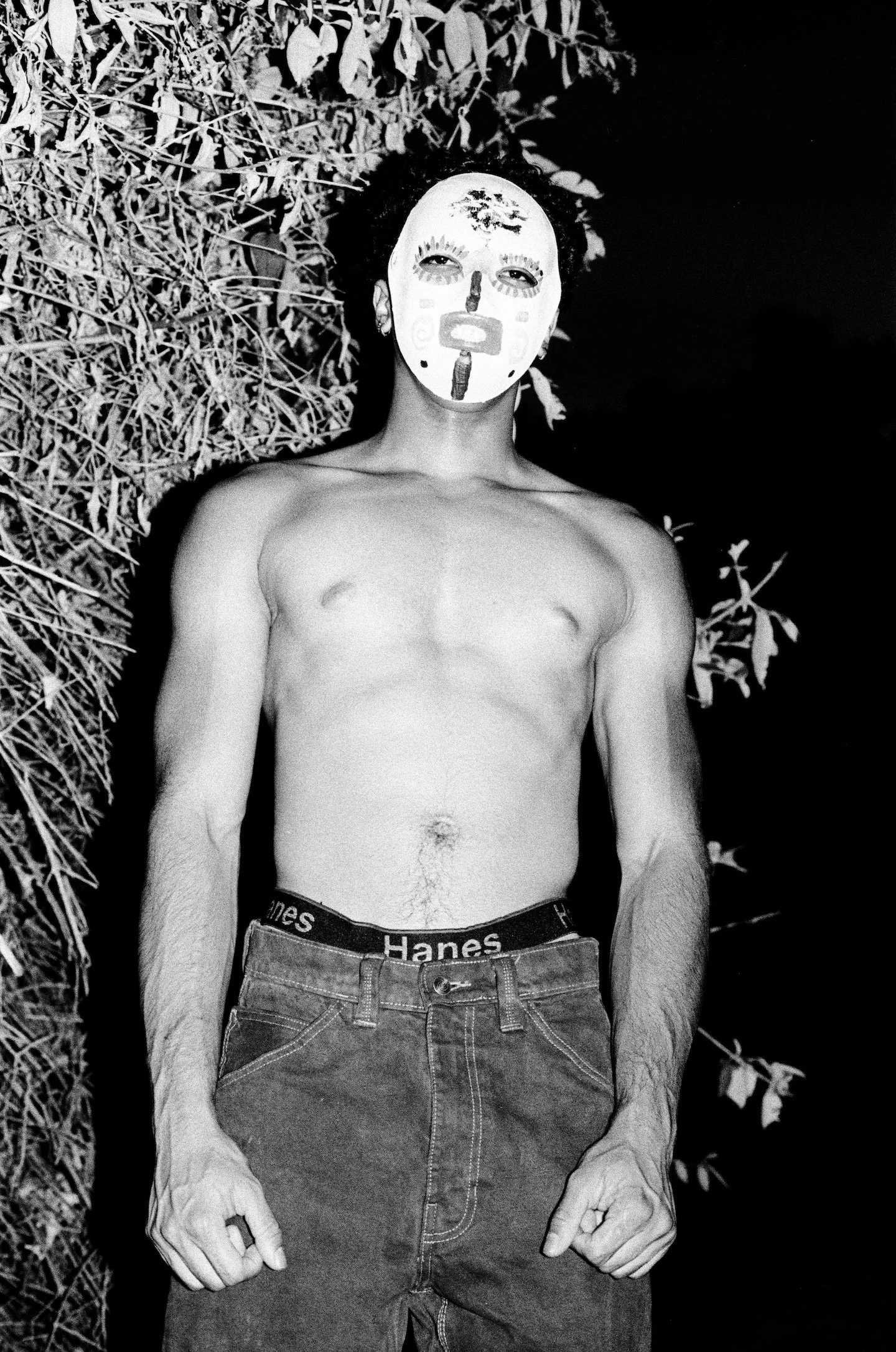 A shirtless young man wearing a painted mask stands outdoors at night with his arms at his sides; direct flash illuminates his body and the dense foliage beside him against a nearly black background.