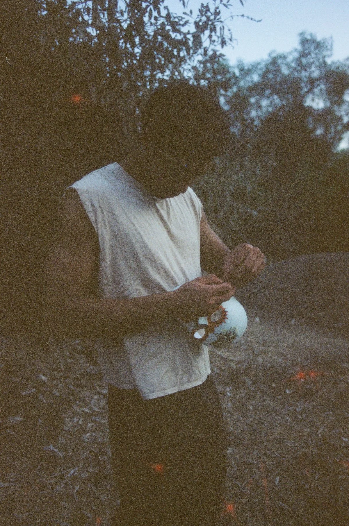 A young man in a sleeveless white shirt stands outdoors at dusk, looking down as he adjusts a painted mask in his hands; the grainy film image is dark, with trees and brush fading into the background.