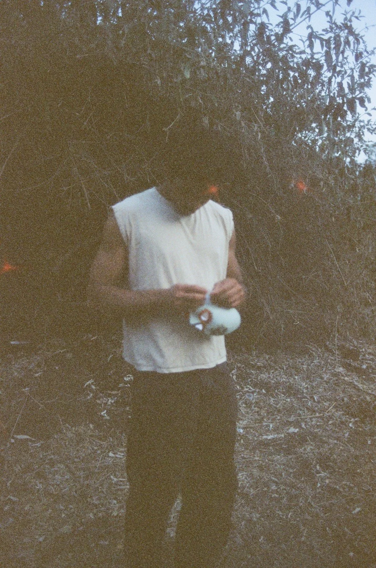 A young man in a sleeveless white shirt stands in a brushy outdoor area at dusk, looking down while holding a a painted mask; the image is soft, grainy, and dim, with dark foliage filling most of the background.