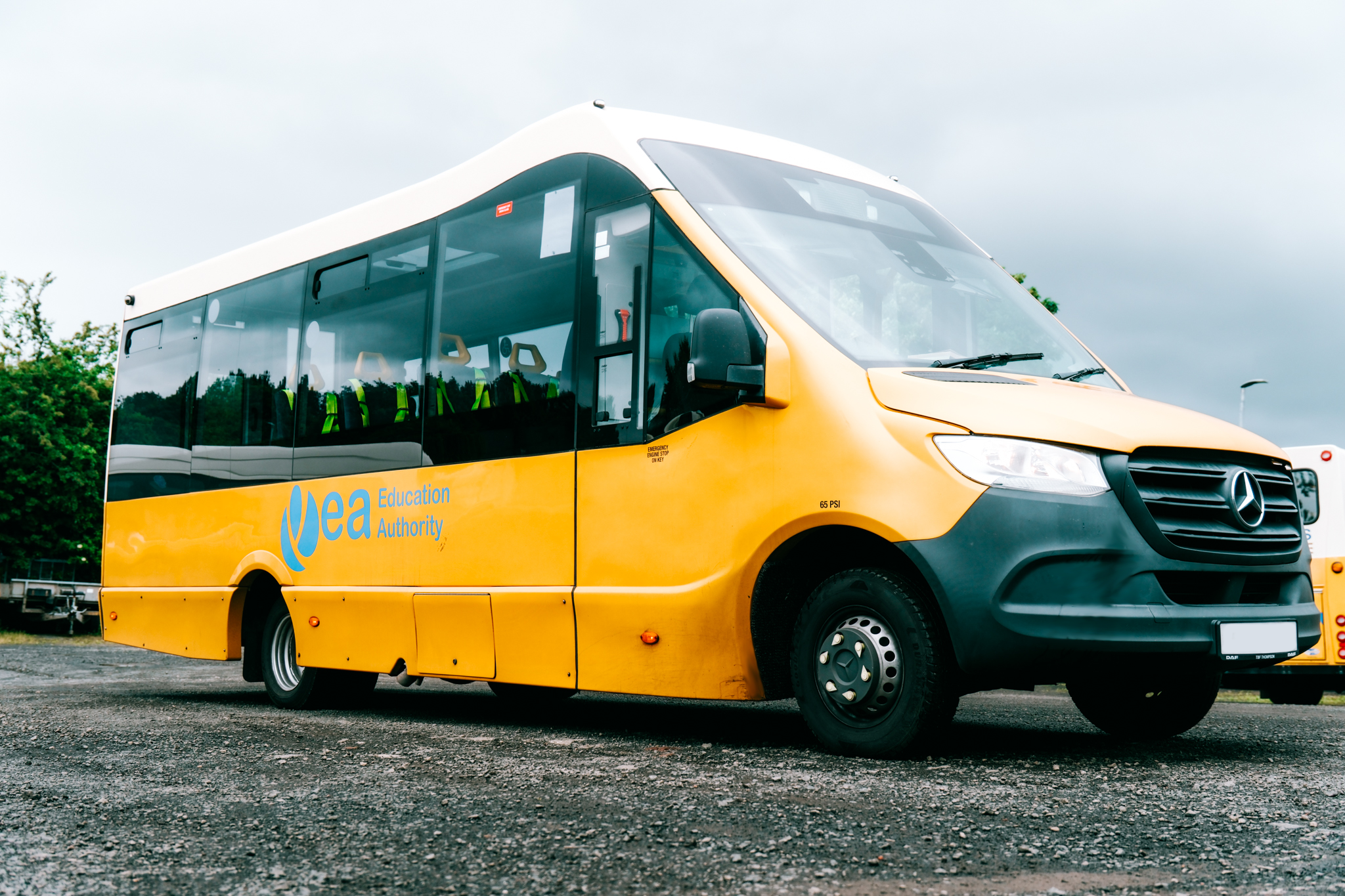 Yellow and black shuttle bus with the logo and text 'EAA Education Authority' on the side, parked on a gravel lot under a cloudy sky.
