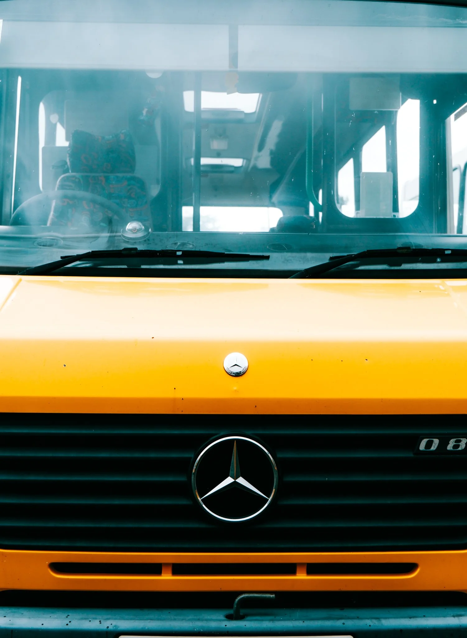 Front view of a Mercedes-Benz vehicle with a yellow hood and a black grille, showing the Mercedes-Benz logo and some model markings, inside a vehicle or garage.